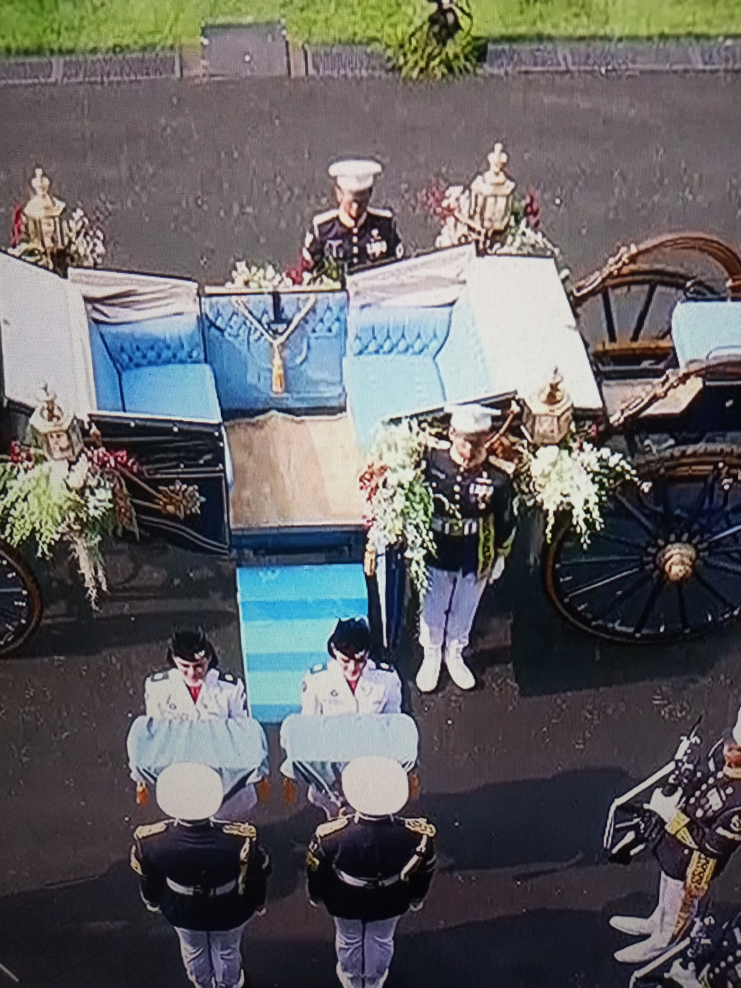 The image shows a solemn procession, likely a funeral, taking place outdoors. In the background, a decorative carriage is visible, adorned with floral arrangements and what appear to be lanterns. The carriage has large wheels and is partially covered with white fabric. Two individuals in military-style uniforms stand near the carriage, one facing left and the other facing right. The individual on the right is wearing a white hat and a dark uniform with medals, and white trousers.
In the foreground, four individuals in similar dark military-style uniforms are positioned in a line. These individuals are carrying what appear to be pallbearer caskets, each draped in a light-colored cloth. Two of these individuals are facing away from the camera, and two are facing towards the camera. The ground appears to be asphalt. The overall atmosphere suggests a formal and respectful event. The specific location in Banyudono, Indonesia is not discernible from the visual information present in the image.