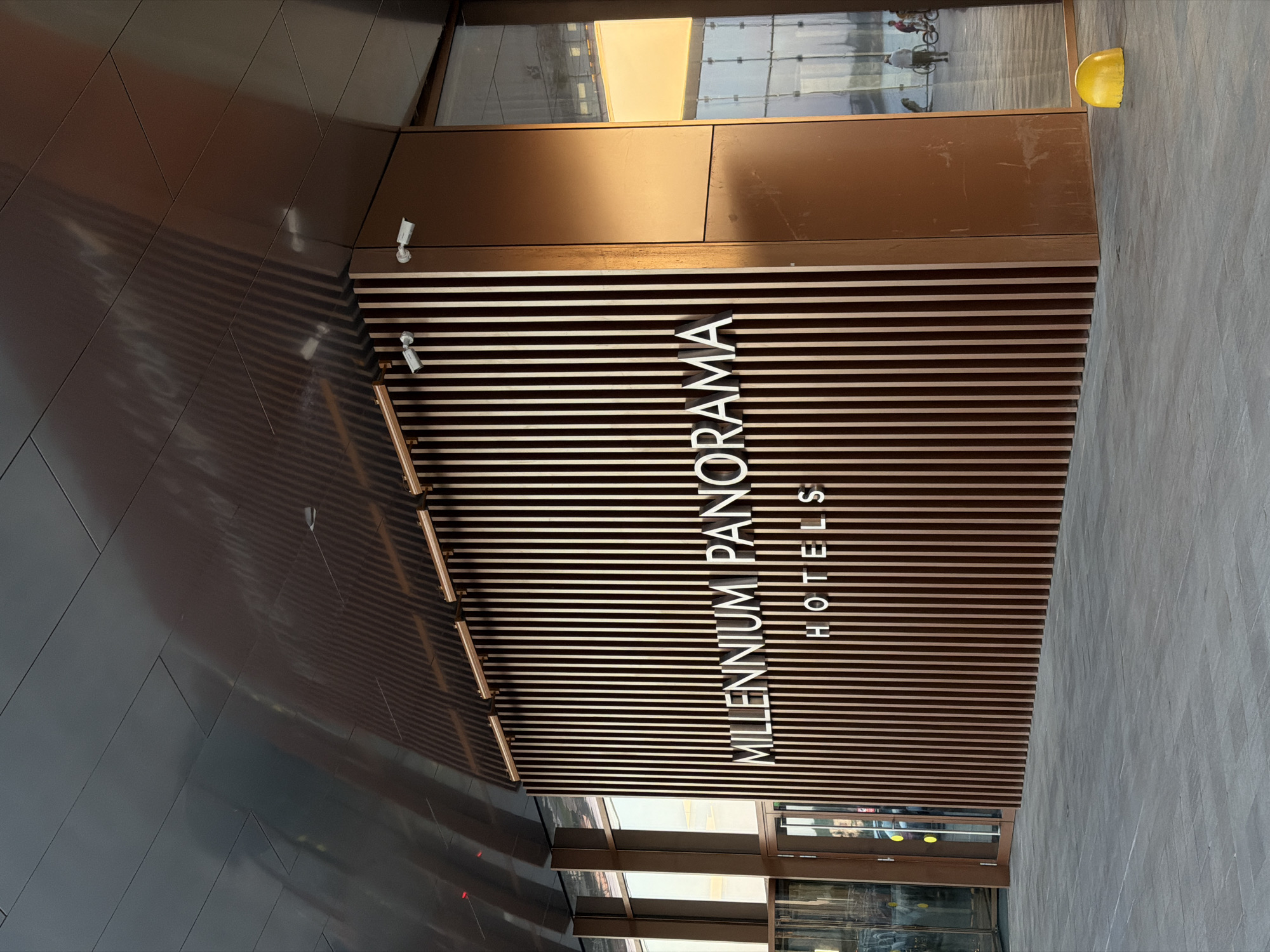 The image shows the entrance of the Millennium Panorama Hotels, located in Kazan, Russia. The hotel name is displayed vertically on a striped brown and white awning. A security camera is visible near the top of the awning. There are people and bicycles visible in the reflection of the glass doors.