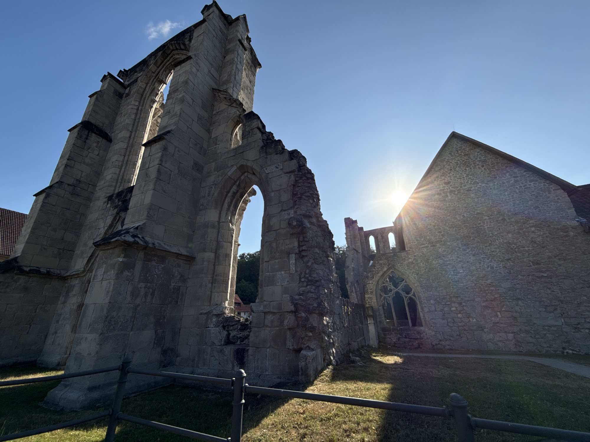 The image displays the ruins of the former Walkenried Abbey in Walkenried, Germany, under a clear blue sky with the sun prominently shining and causing lens flare. The foreground features a black metal fence running horizontally across the frame. Beyond the fence, the ground is covered with dry grass and patches of sunlight.
The central subject is the stone architecture of the abbey ruins. On the left, a large, weathered stone structure with an arched opening dominates the frame, showing evidence of decay and missing sections of stonework. The texture of the stones is rough and aged. To the right of this main structure, another section of the ruins is visible, including a prominent arched window with intricate stone tracery. The building materials are primarily large, rough-hewn stones.
In the distance, on the right side of the image, a section of the abbey appears to be more intact, with a sloped stone roof and a gabled end. The sun is positioned behind this structure, creating a bright halo and radiating light rays that extend across the sky and onto the ruins. A faint trace of a building with a red roof is visible in the background, behind the left-hand ruins. No people are present in the image. The overall impression is one of historical grandeur and the passage of time.