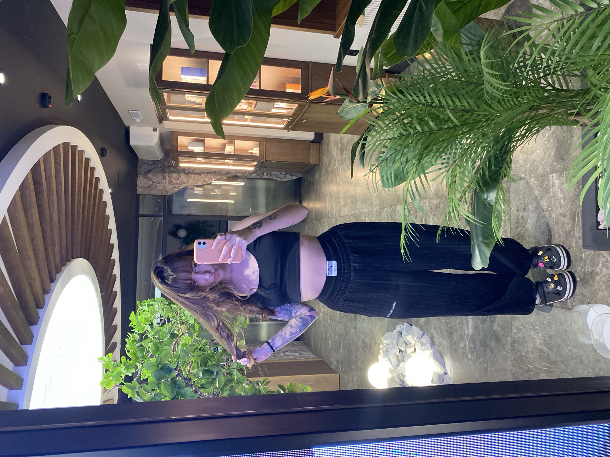 A woman is taking a mirror selfie indoors. She is wearing a black crop top and black pants. She has tattoos on her arm. The background includes plants, a decorative wooden arch, and display cases. The location appears to be a shop or a business in an urban area. The city of Votkinsk, Russia, is not discernible from the image.