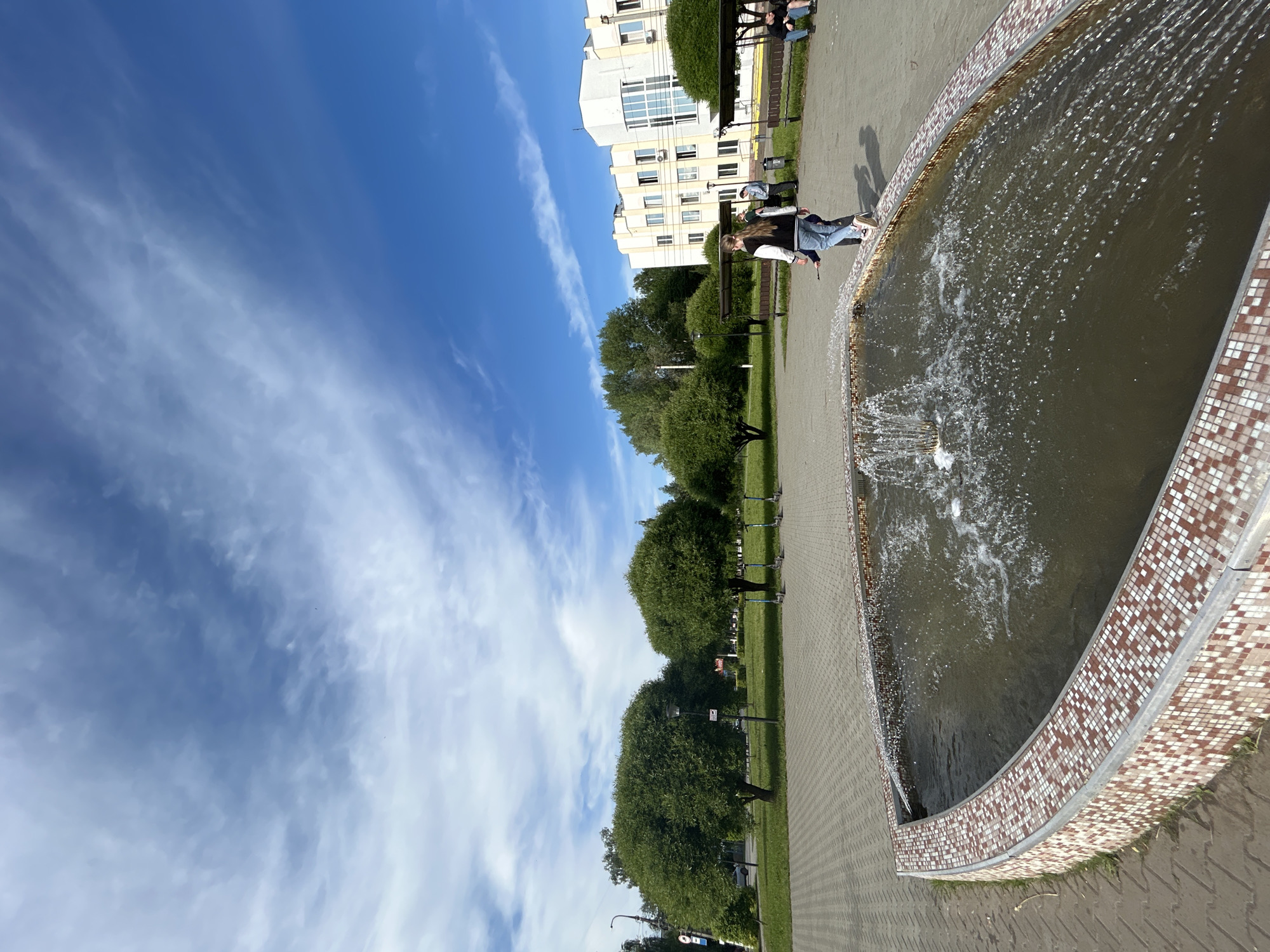 This image captures a vibrant outdoor scene in Chaikovsky, Russia, on a clear, sunny day. The main subject is a large, crescent-shaped fountain actively spraying water, its edge decorated with distinctive brown and white mosaic tiles. The water within the fountain appears somewhat murky.
To the right of the fountain, a wide paved walkway extends into the distance, flanked by lush green grass and numerous densely leafed trees, creating a park-like setting. Several dark-colored park benches are placed along the path and within the grassy areas. In the middle distance, a line of electric scooters is parked on the grass, suggesting public transport options.
In the background, a modern, multi-story building with white and gray facades and many windows is visible, indicating the urban context of the park. Several people are present, enjoying the pleasant weather; one person is seated on a bench, and another is standing near the fountain. Long shadows on the paved ground suggest it is either morning or late afternoon. The sky is a brilliant blue with wispy, streaky cirrus clouds, indicative of fair weather. The scene conveys a calm and leisurely atmosphere in a public recreational area. While some signs are visible on poles, any text they might contain is illegible.