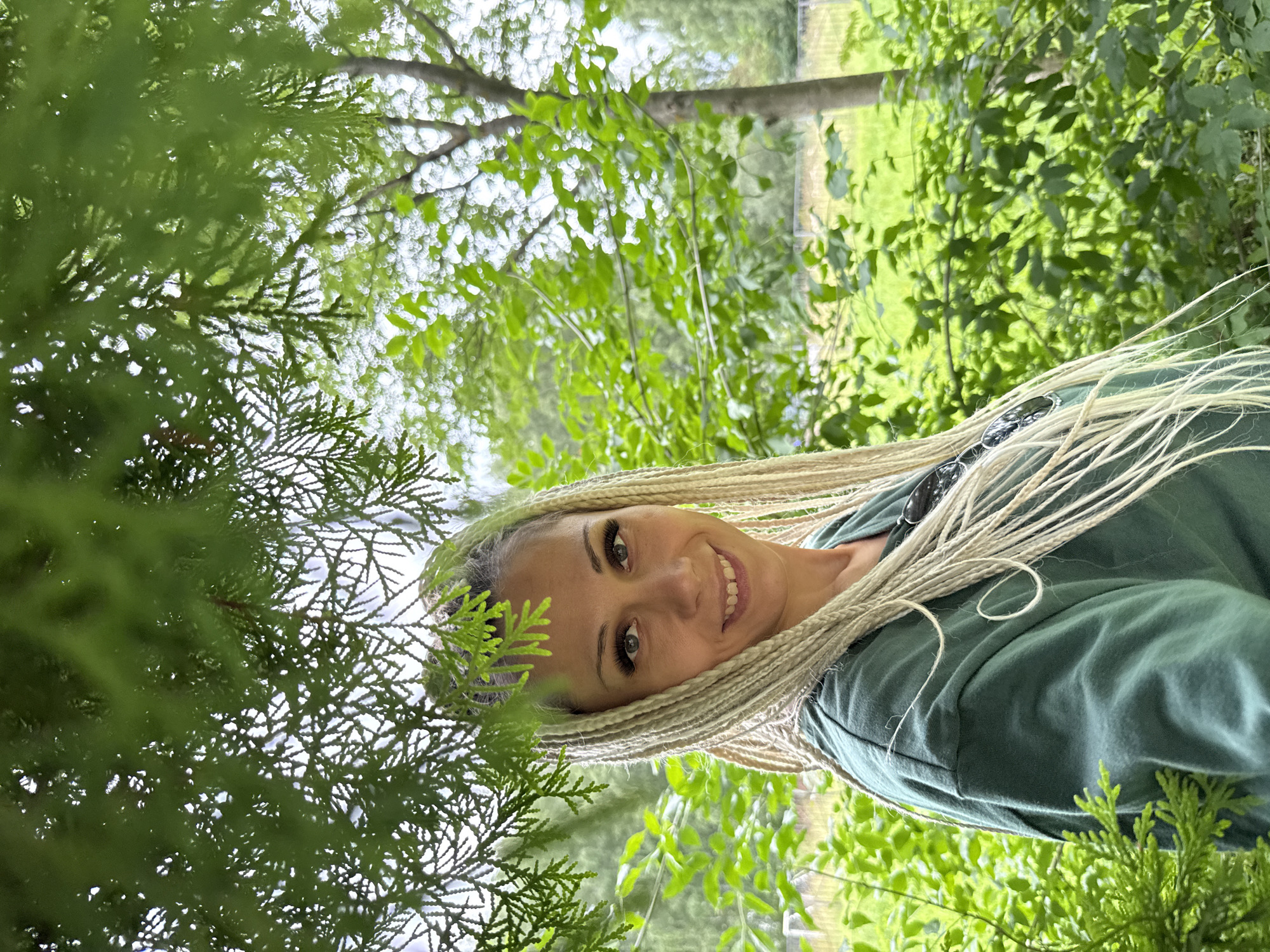 A woman with blonde braided hair and dark eye makeup is smiling and looking at the camera, partially obscured by green foliage. She is wearing a dark green sweatshirt. The background consists of lush green trees and grass, suggesting an outdoor, possibly park-like setting. The image appears to have been taken in Rusanovo, Russia.