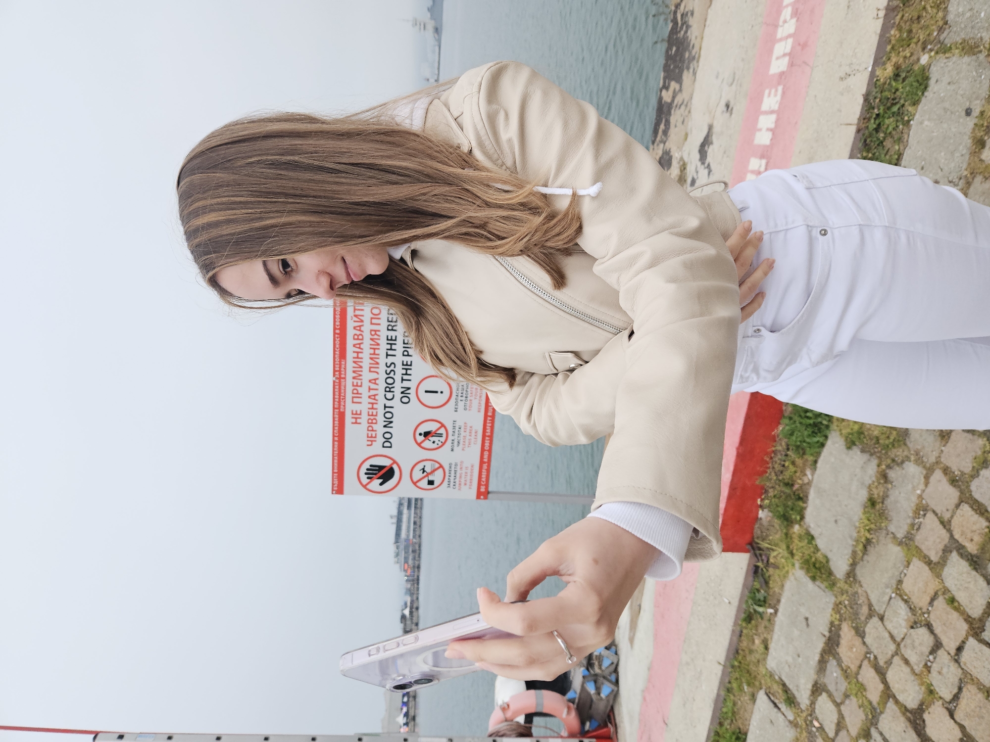 A young woman with long brown hair, wearing a beige jacket and white pants, is standing on what appears to be a pier or waterfront area in Varna, Bulgaria. She is holding a smartphone, possibly taking a photo or video. A sign nearby displays warnings in Bulgarian and English ("Do Not Cross the Red Line on the Pier") and safety rules for the harbor. The background includes the sea and a somewhat cloudy sky. The ground is made of stone and concrete with patches of grass.