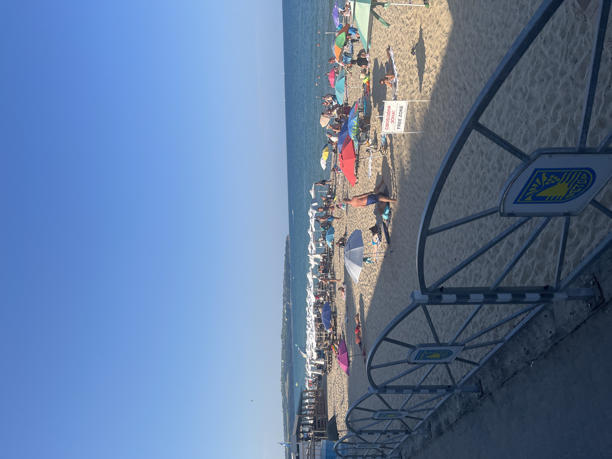 The image depicts a crowded beach in Obzor, Bulgaria, on a sunny day. The foreground features a metal fence with blue and yellow emblems. Beyond the fence, a sandy beach is filled with numerous beach umbrellas of various colors, providing shade for sunbathers. Several people are visible lounging on towels and chairs, some in swimwear, while others are walking or standing. The sea, a calm blue, stretches to the horizon, with a few boats in the distance. A sign that reads "СВОБОДНА ЗОНА! FREE ZONE" is prominently displayed on the sand. A building or structure with a covered area and tables is situated on the left side of the beach, likely a restaurant or bar. The overall scene conveys a typical summer vacation atmosphere.