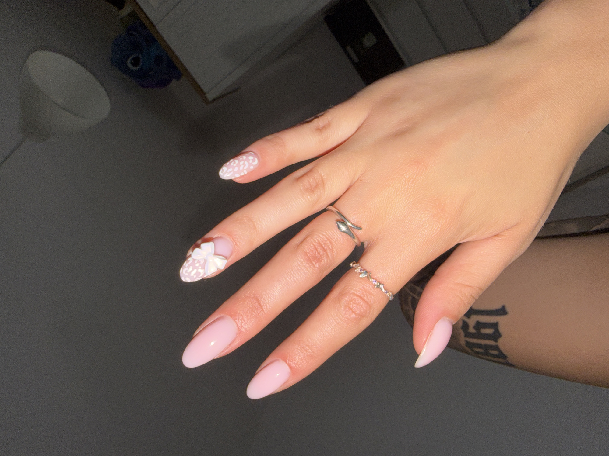 A left hand is positioned in the foreground, palm slightly turned inward, showcasing manicured nails. The skin tone is light to medium. The fingernails are of an almond shape. The pinky, ring, and middle fingers display a solid light pink nail polish. The index finger features a light pink base with a white, abstract, or leopard print pattern, and a three-dimensional white bow embellishment is affixed near the cuticle. A silver ring with small, multi-colored, leaf-shaped stones is present on the ring finger. Another silver ring with a wavy or snake-like design is on the middle finger. A portion of the thumb's nail, also light pink, is visible in the lower-left corner. Below and to the left of the hand, a tattoo in black, old English-style script shows the numbers "861" on an arm or shoulder. The background consists of a plain dark gray wall. In the upper right quadrant, a white conical pendant light fixture hangs from a dark ceiling. Below the light fixture, on a white shelf or furniture item, is a dark blue plush toy with large eyes, and additional white furniture is partially visible to the right. The indoor setting provides no specific geographical indicators for Plovdiv, Bulgaria.