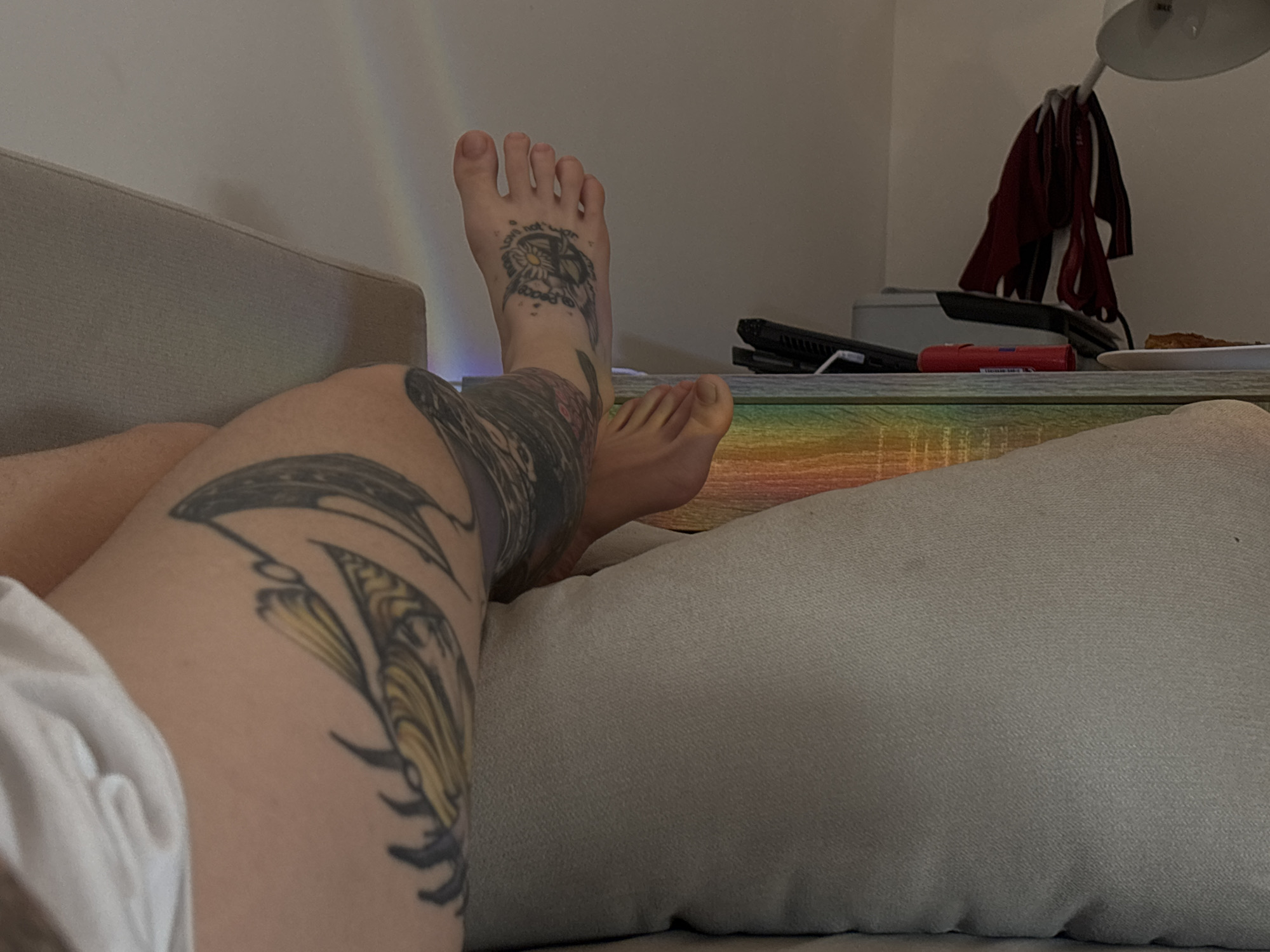 This image depicts an indoor scene, primarily focusing on the lower limbs of an individual resting on a light-colored couch or cushion. The image does not contain information to determine its specific location in Zvezdara Urban Municipality, Serbia.
The subject's left leg, visible in the foreground, features an extensive tattoo covering the thigh and calf. This tattoo consists of intricate black and grey outlines with notable yellow and gold shading, forming abstract or organic shapes, possibly resembling wings or stylized leaves. The left foot also has a tattoo on its dorsal surface, featuring a peace symbol centrally, encircled by the text "love not war," and a daisy-like flower design. The subject's right foot is bare and appears free of tattoos. White fabric, likely shorts or undergarments, is visible on the upper left, partially covering the upper left thigh.
To the right of the subject's feet, a wooden-patterned surface, possibly a desk or table, is visible. This surface exhibits a colorful, gradient reflection spanning the visible spectrum (rainbow colors), suggesting the presence of an LED light strip positioned beneath it. Objects on this surface include a black electronic device, possibly a laptop or game console, along with associated cables. A red cylindrical object, possibly a marker or lighter, is also present. Further right, a white plate holds a single slice of food, resembling pizza or a similar baked item. Above this table, a white flexible-arm desk lamp is attached. Several pieces of dark red fabric, appearing to be undergarments or small clothing items, are hanging from the lamp's arm. The background consists of a plain, light-colored wall. The overall lighting is subdued, suggesting an indoor evening or dimly lit environment.