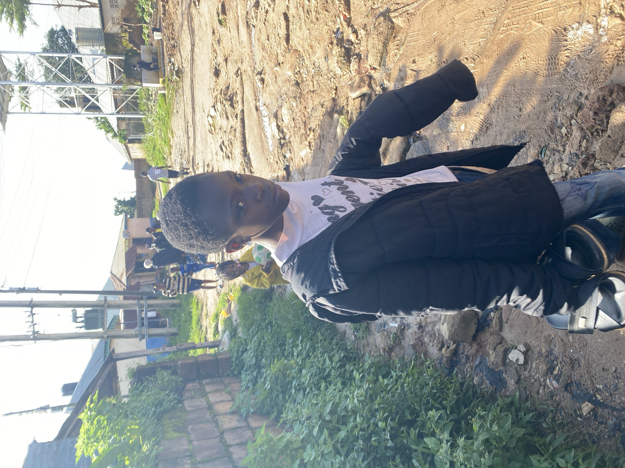 This media file features a young person in the foreground, wearing a dark puffer jacket over a white t-shirt with a heart and text design. They are looking directly at the camera with a neutral expression. The setting appears to be a somewhat rural or undeveloped area in Barakin Lerawi, Nigeria. Behind the main subject, other people and buildings are visible, suggesting a community or village environment. In the background, there are structures that look like homes, some with metal roofs, and a tall metal tower that could be a communication mast or part of a water system. The ground is a mixture of dirt, gravel, and some patches of greenery. Sunlight suggests it is daytime, likely in the morning or late afternoon due to the quality of the light. The overall atmosphere is casual, with individuals going about their day. The presence of electrical poles and what looks like a transformer indicates infrastructure.