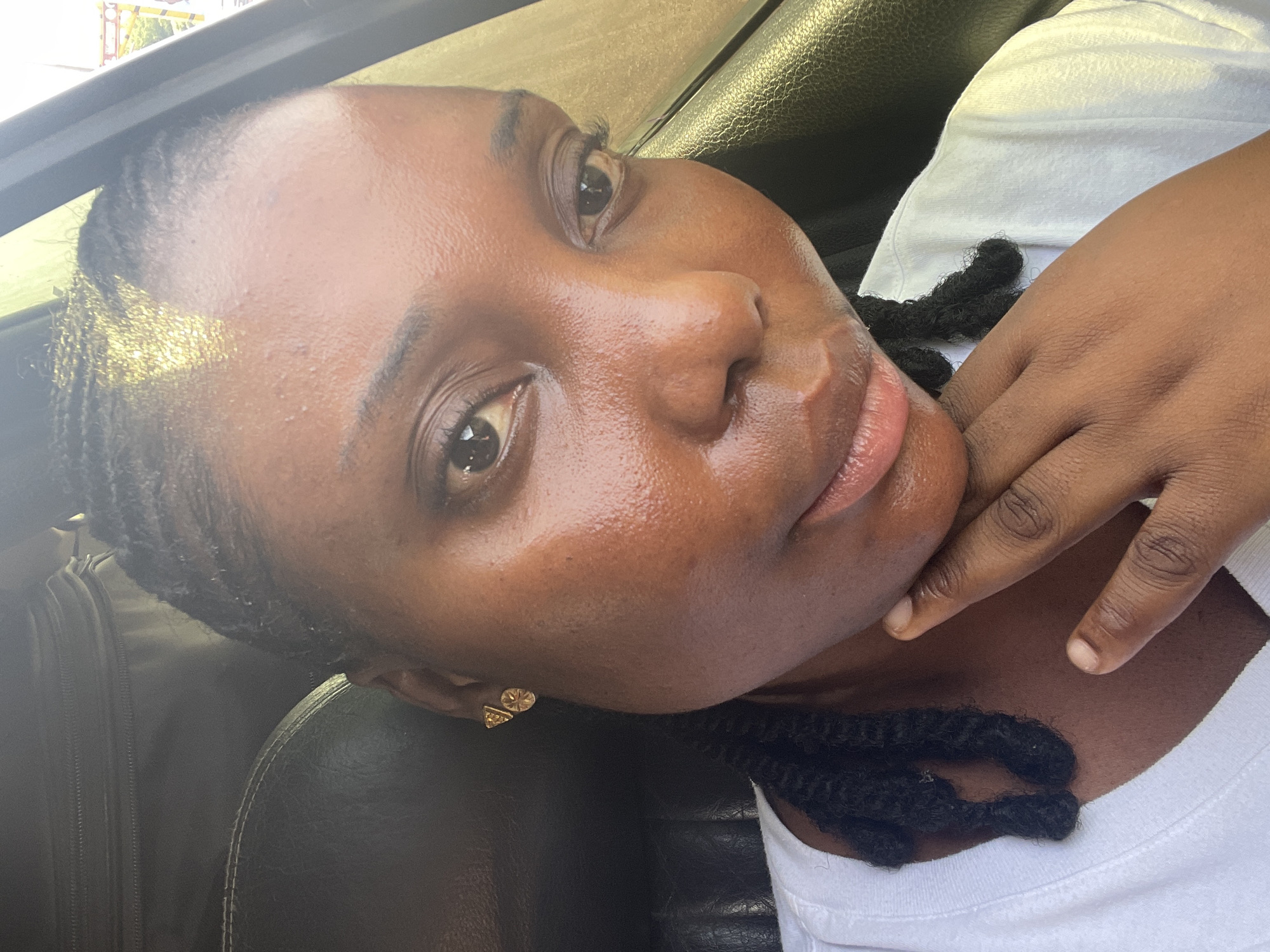 A close-up, slightly tilted portrait captures a person with dark skin, likely female, inside a vehicle during the daytime in Keffi, Nigeria. The subject looks directly at the camera with a calm, relaxed expression and a subtle smile. Her skin appears dewy or oily, reflecting the bright natural light.
Her dark hair is styled in braids or twists, with strands visible against the white crew-neck t-shirt she is wearing. Small gold-colored stud earrings adorn her right ear; one is a triangle, and the other appears to be a flower or star shape. One hand is casually placed near her chin and neck.
The background is softly out of focus, revealing the dark upholstered interior of what appears to be a car, including a seat and a glimpse of bright light from outside a window. The clear lighting suggests a sunny day. No text is visible in the image.