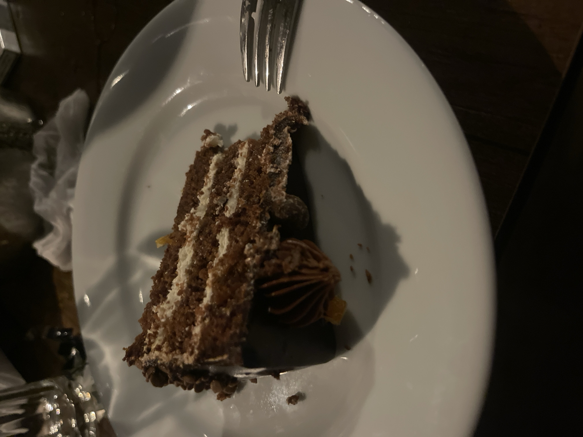 A slice of chocolate cake with layers of frosting and chocolate glaze sits on a white plate. A fork is positioned to the right of the cake. The scene is dimly lit, with the plate resting on a dark wooden surface. There is no indication of the city of Ruse, Bulgaria.