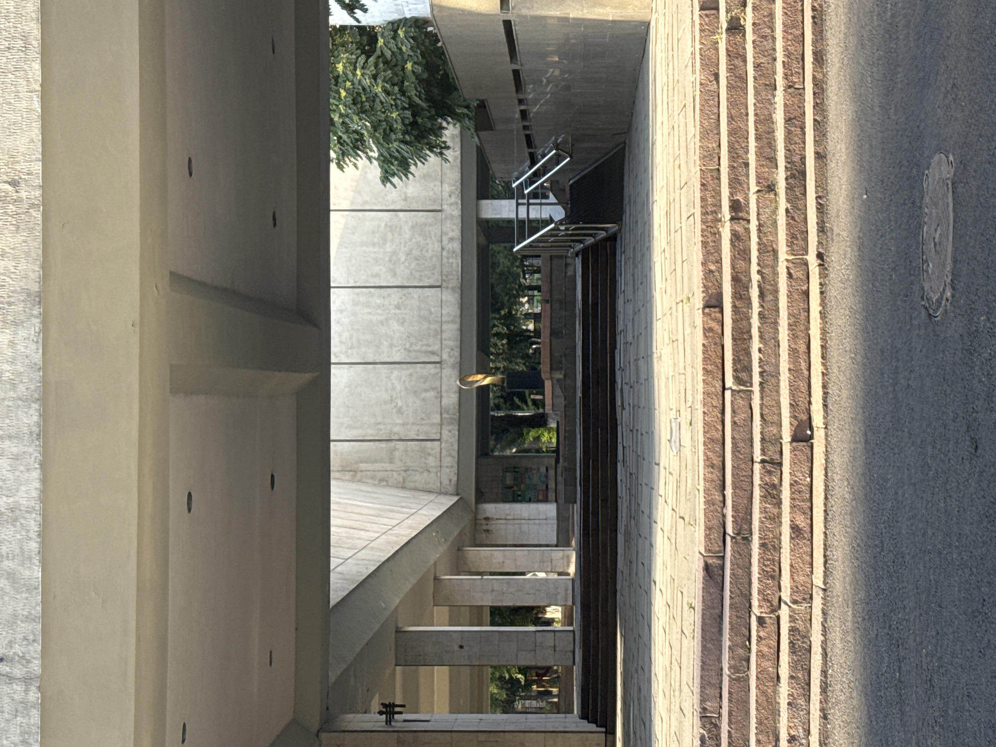 The image depicts a wide shot of the exterior entrance to the State Historical Museum in Bishkek, Kyrgyzstan. The perspective is from ground level, looking up towards the museum's entrance, which is framed by a large, overhanging concrete structure.
In the foreground, there are several steps made of rough-hewn reddish-brown stone. These steps lead up to a large, paved plaza area, also constructed of stone slabs. The plaza extends under the overhang and into the museum's entrance area.
The entrance itself features a colonnade of tall, square concrete pillars. Beyond the pillars, the museum's interior is visible, with a large open space and further architectural elements. A golden sculpture, possibly abstract or figurative, is visible in the background, standing on a pedestal.
To the right of the main entrance, there is a metal railing and a ramp leading up to a higher level, suggesting an alternative access point. A portion of a large green tree with dense foliage is visible in the upper right corner, contrasting with the predominantly concrete and stone architecture.
The lighting suggests it is either early morning or late afternoon, with long shadows cast across the plaza and steps, indicating the sun is at a low angle. The overall scene is one of monumental architecture, with a sense of scale and grandeur. No people are clearly visible in the frame.