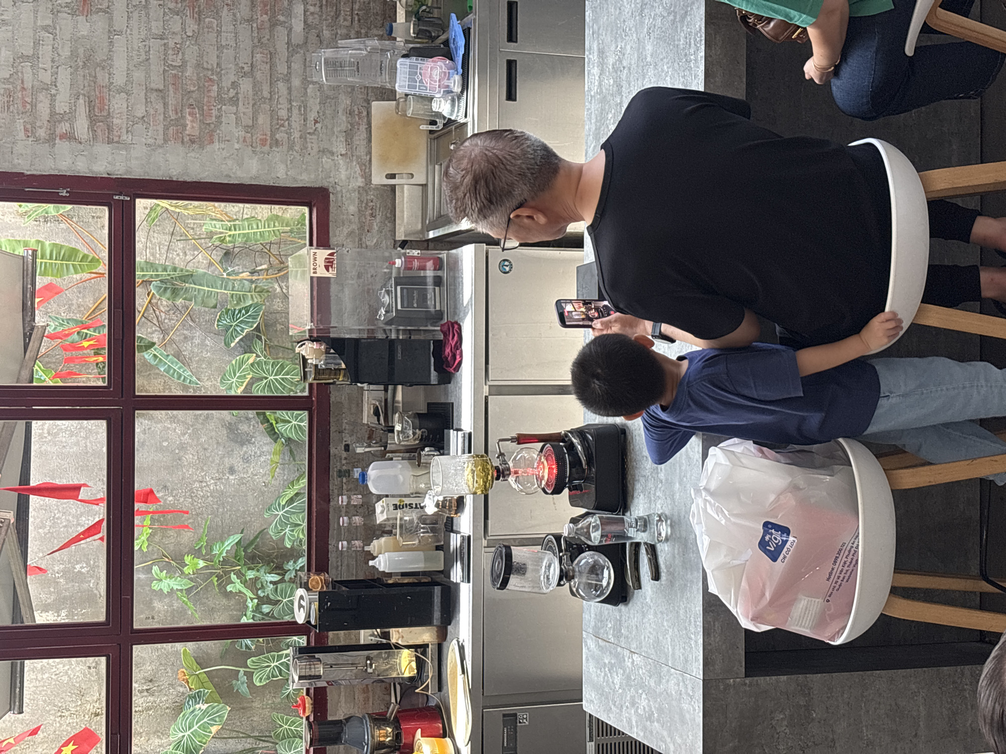 The image shows a view from behind a man and a boy sitting at a counter in what appears to be a coffee shop or cafe. The man, wearing a black t-shirt and glasses, is holding up a smartphone, possibly taking a picture or recording a video of a coffee brewing apparatus on the counter. The boy, in a blue t-shirt, stands next to him, also looking at the phone.
In the background, a large window overlooks an outdoor area with lush green plants and red flags hanging. The interior of the cafe features a brick wall, shelves with various bottles and containers, and kitchen appliances like a blender. A siphon coffee maker is prominently displayed on the counter, with its glass bulbs and heating element.
The counter itself is made of a grey, speckled material, and a large white bag containing pink items sits on a white stool in the foreground. The overall setting suggests a modern, well-lit cafe environment. The presence of the red flags outside could indicate a cultural celebration or event in Đà Nẵng, Vietnam, a coastal city known for its vibrant atmosphere and numerous cafes.