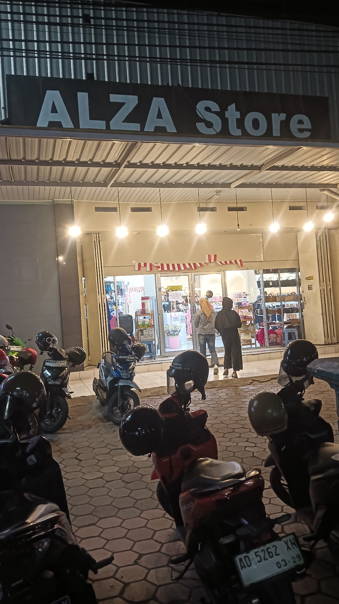 The image shows the exterior of a store named "ALZA Store" in Sragen, Indonesia. The store is illuminated by overhead lights and appears to be open, with people visible inside. In front of the store, a row of motorcycles is parked on a paved area. The store has a modern facade with large glass doors and windows. The name "ALZA Store" is prominently displayed in white lettering on a dark background above the entrance. Indonesian flags, in red and white, are displayed horizontally across the top of the store entrance. Inside the store, shelves are stocked with various items, and at least two individuals are seen browsing. The overall scene suggests a commercial establishment operating in the evening.