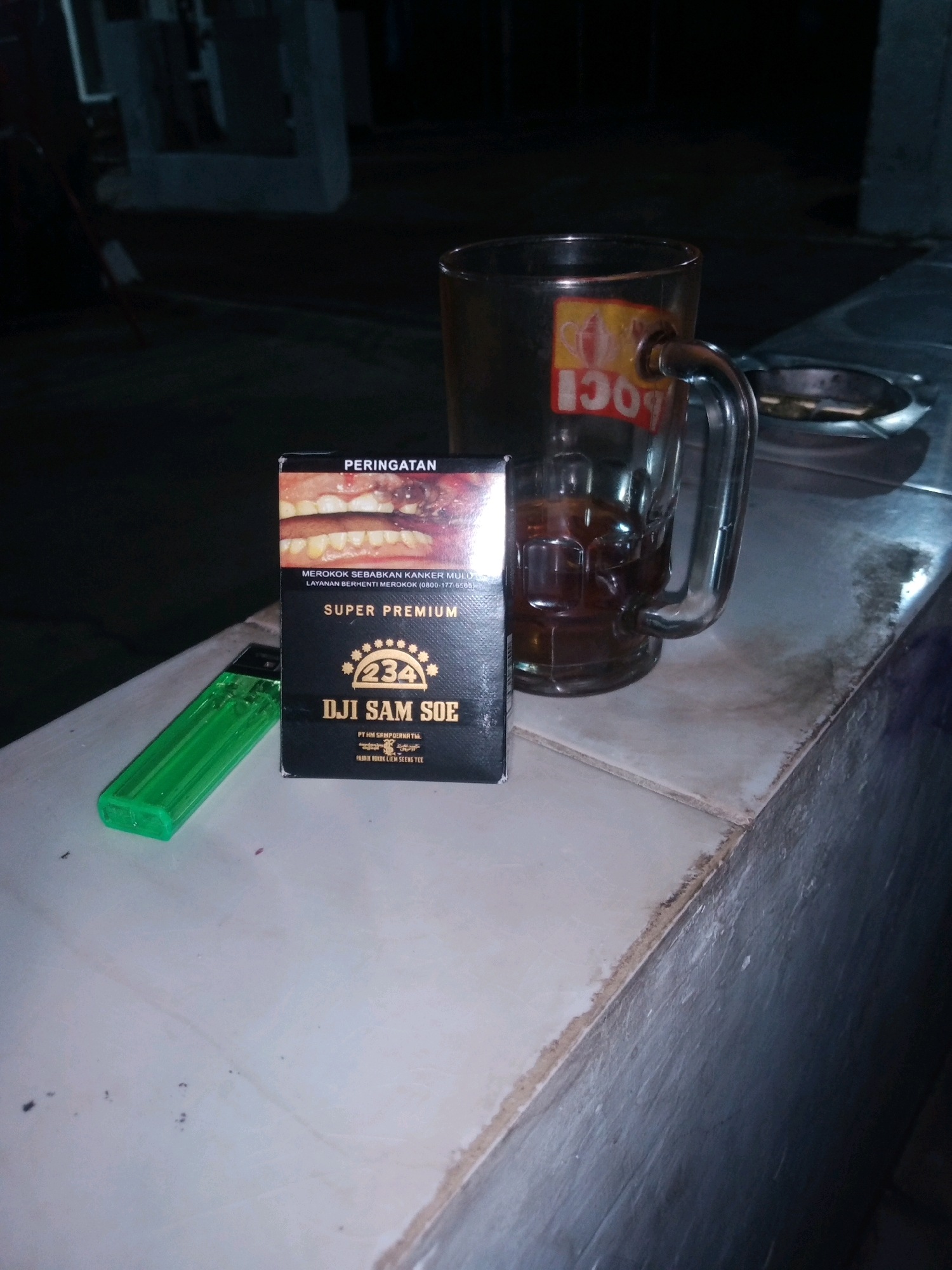 In Rejasari, Indonesia, a close-up shot captures a cigarette pack and a green lighter resting on a white tiled surface. The cigarette pack, labeled "DJI SAM SOE SUPER PREMIUM," features a graphic warning of diseased teeth. Beside it, a glass mug filled with a dark beverage stands, its side adorned with a sticker bearing the "POCI" logo. An ashtray, partially visible, suggests a recent or intended smoking activity. The background is a dark, indistinct urban setting, hinting at an outdoor, possibly nighttime environment.