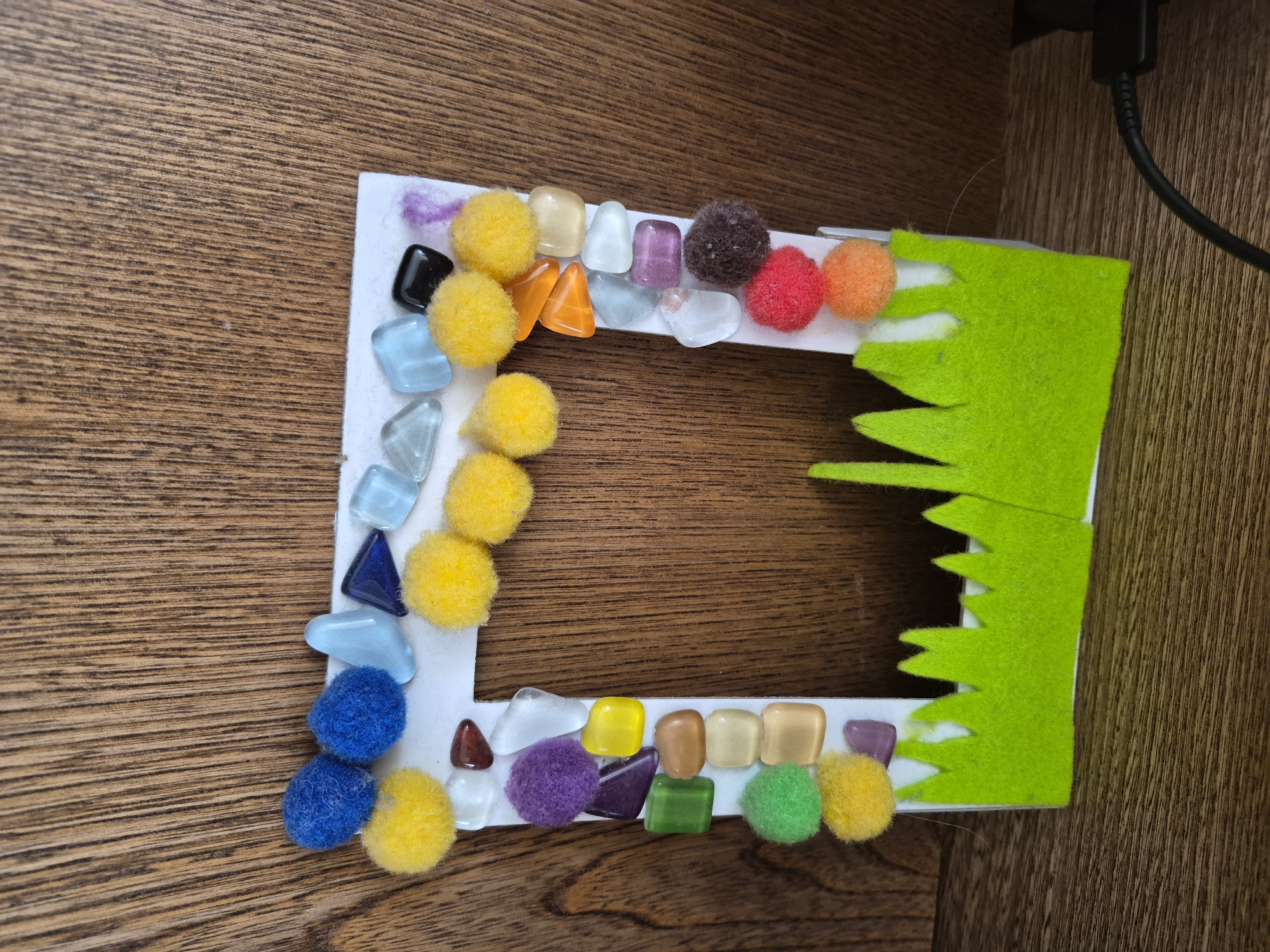 This is a close-up, eye-level, indoor shot of a handmade picture frame against a wooden background. The frame is white with a rectangular opening and is decorated with various colorful craft supplies. Along the top edge, there are blue and yellow pom-poms interspersed with light blue and dark blue triangular glass pieces and a black square. The left side of the frame features a vertical line of glass gems in various shapes and colors including purple, yellow, green, and tan, with interspersed blue and yellow pom-poms. The right side of the frame continues the decoration with more glass gems in orange, light blue, and purple, and pom-poms in brown, red, and orange. The bottom of the frame is adorned with a scalloped green felt strip resembling grass. The wooden background has a prominent grain pattern. The overall impression is a whimsical and colorful craft project. The scene is located in Unknown, Unknown.