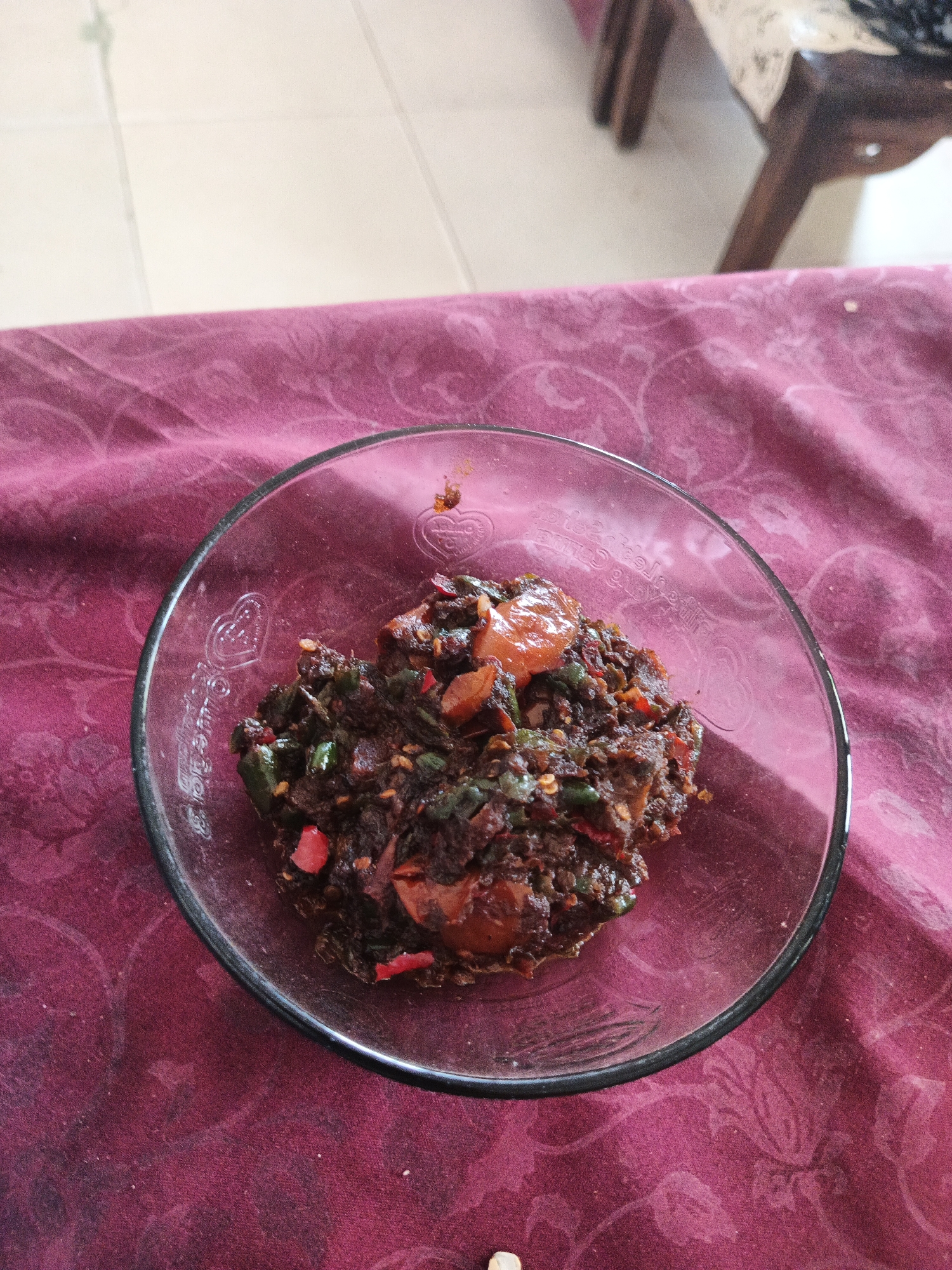 A close-up, overhead shot shows a clear glass bowl filled with a dark, chunky mixture of food, resting on a reddish-purple tablecloth. The food appears to be a stir-fry or a condiment, with visible pieces of red chili, green chilies, and possibly onions. The bowl has intricate embossed designs, including hearts, along its rim. The tablecloth features a subtle floral pattern. In the background, out of focus, are light-colored tiled flooring and the legs of a wooden chair. The scene is indoors and the lighting suggests a daytime setting. The specific location in Rejo Basuki, Indonesia, cannot be determined from the visual information provided.