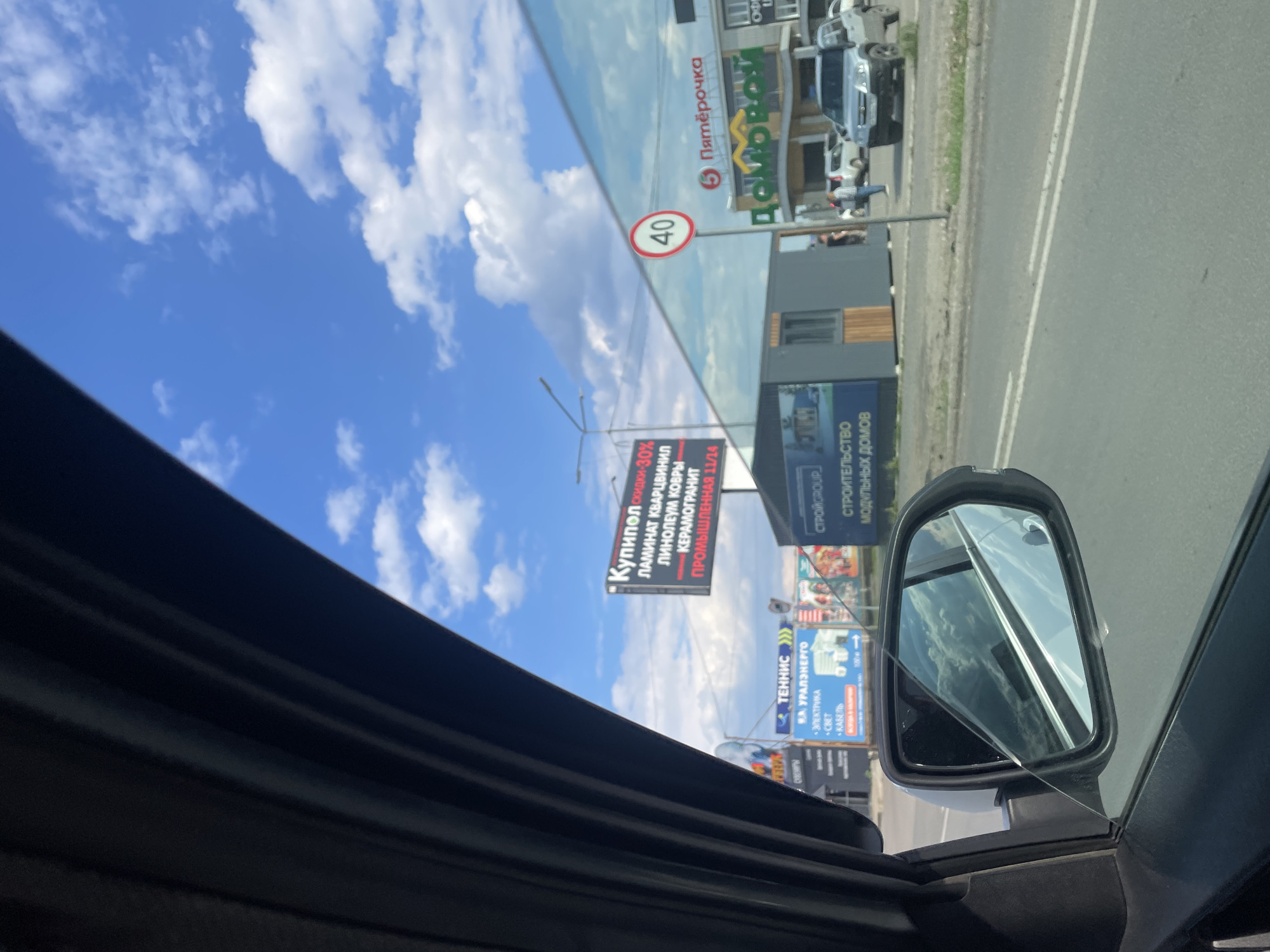 The image captures a street scene through the window of a vehicle in Chaikovsky, Russia, on a sunny day with scattered clouds. In the foreground, the side mirror of the car reflects the sky and clouds. To the right, a modern building with a reflective facade dominates the background. Prominently displayed on this building is the logo for "Пятёрочка," a Russian supermarket chain, and another sign for "ДомОвой," likely a home goods store. A speed limit sign indicating "40" is visible. In front of the building, a small portable kiosk or booth is positioned on the roadside. Several vehicles are parked or driving on the road. Further to the left, various billboards are present, including one advertising "Теннис" and another with Russian text related to building materials and services, and a third billboard with text "КУпипол" and "скидки 30%". The overall environment suggests a commercial or mixed-use area within the city.