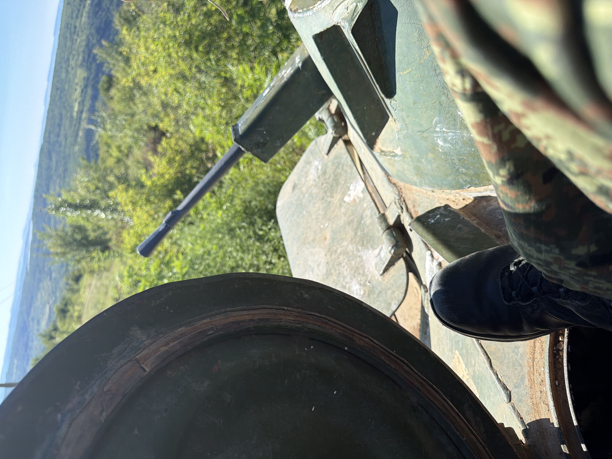 A close-up, eye-level, outdoor shot features the interior of a military vehicle, possibly a tank, with the open hatch revealing a lush, green, hilly landscape in the background. In the foreground, a person's black combat boot is visible, resting on the vehicle's metal floor. Part of the vehicle's turret, including a portion of a gun barrel, is also in view. The person is wearing camouflage trousers, partially obscuring the view. The lighting suggests it is daytime, with a clear, blue sky overhead. The location is Beliakovets, Bulgaria.