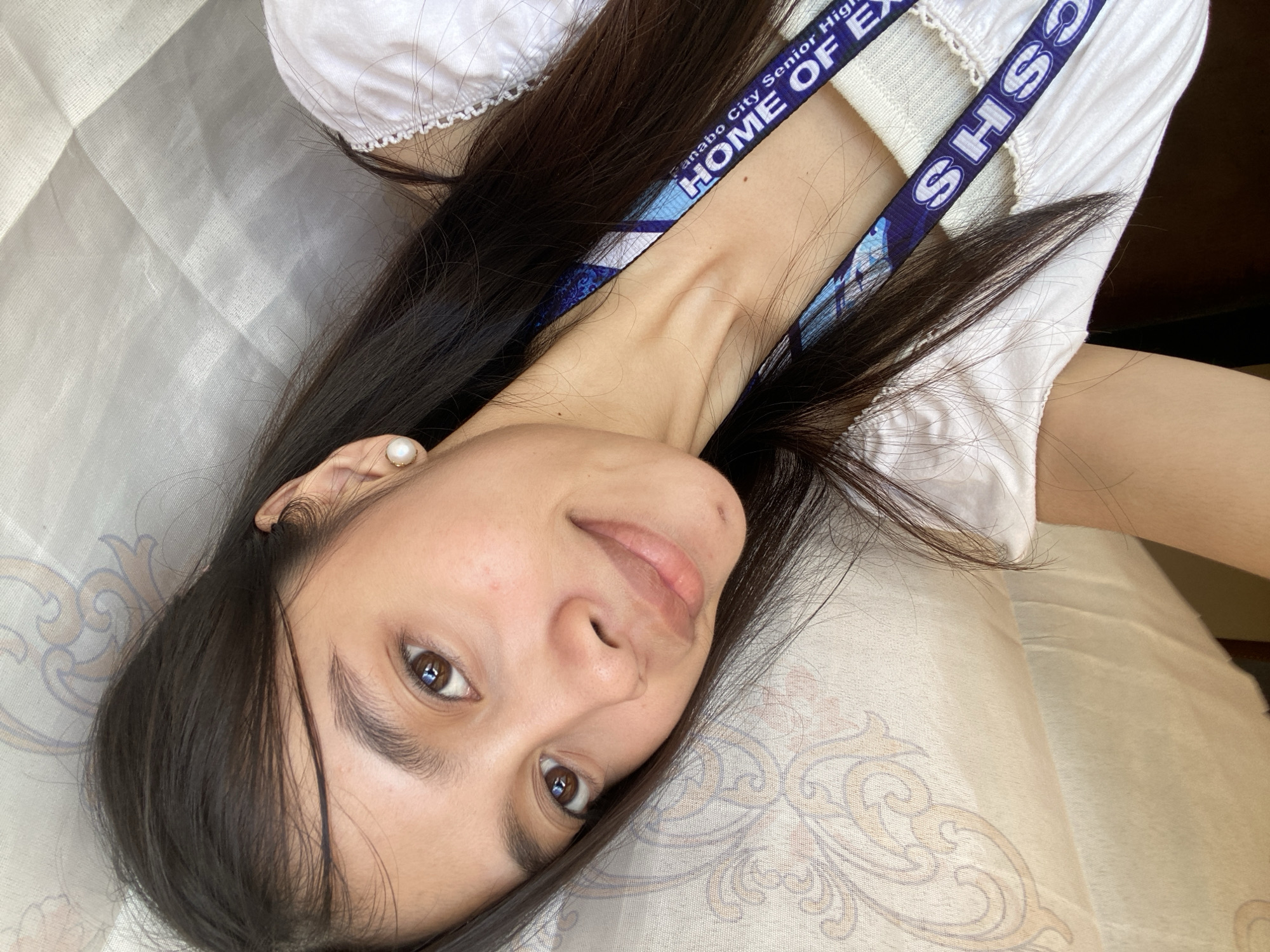 In a medium shot, a young woman with long, dark hair and brown eyes poses for a selfie. She is wearing a white top with a ruffled neckline and a lanyard with the text "CSHS" and "HOME OF EXCELSIOR" which appears to be related to the Panabo City Senior High School in Panabo City, Philippines. She has a small pearl earring in her right ear and a mole on her left cheek. The background consists of a light-colored, textured curtain with a subtle ornate pattern. The lighting suggests it is daytime, possibly near a window.
