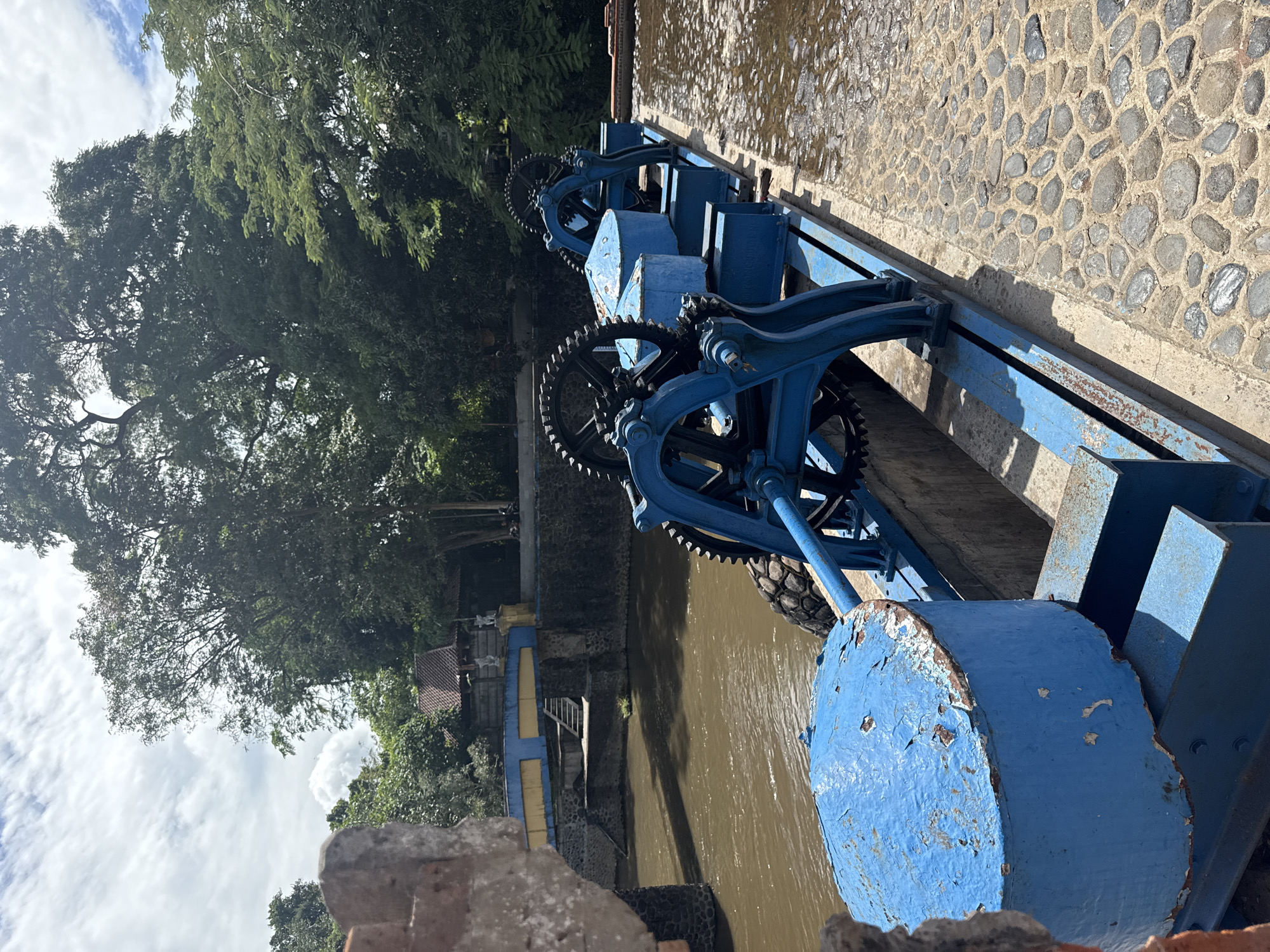 The image shows a blue-painted water dam mechanism with large gears and cylindrical supports situated on a stone embankment, presumably along a river. The water below the dam is murky brown. In the background, there's a cluster of trees and a glimpse of a building with blue and yellow accents. Based on the location provided, the image depicts the Tukad Bindu Dam in Denpasar, Indonesia.
