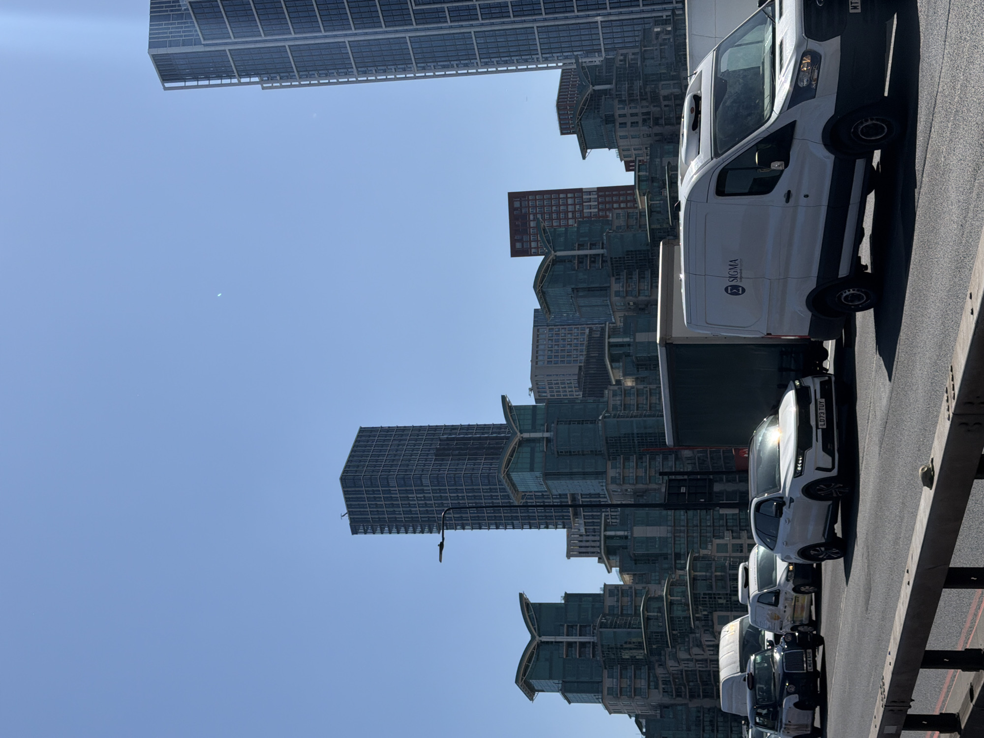 The image depicts a traffic scene in London, United Kingdom. The view is from an elevated position, possibly a bridge or overpass, looking down at a line of stationary or slow-moving vehicles, including a white van marked "SIGMA," several cars, and a London taxi. The background features modern high-rise buildings with glass facades, suggesting a contemporary urban environment. The sky is clear and blue, indicating a sunny day. The focus is primarily on the vehicles and the urban backdrop.