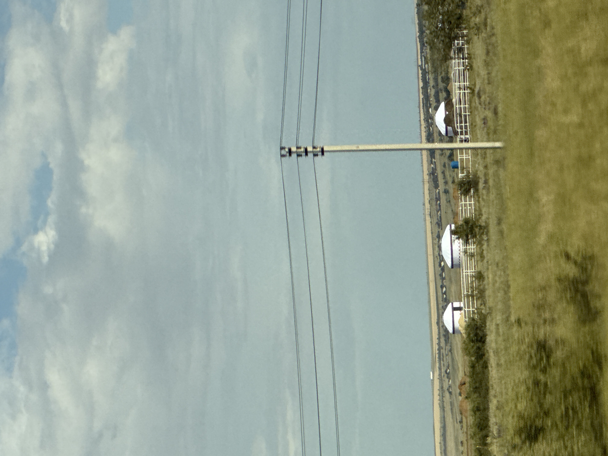The image depicts a pastoral scene in Дорожный, Russia, characterized by a vast, open sky filled with scattered clouds, suggesting a daytime setting. A prominent power line stretches across the frame, supported by a white pole with dark insulators, indicating the presence of electrical infrastructure. In the background, a grassy field unfolds, dotted with small white structures that appear to be shelters or perhaps part of a farm or outdoor facility. These structures have a distinctive domed roof design. A thin line of trees or brush marks the transition between the field and a distant, pale blue expanse, possibly representing water or a hazy horizon. The overall impression is one of rural tranquility and openness, with the power lines providing a contrasting man-made element against the natural landscape. No people are visible in the image, and there is no discernible text.