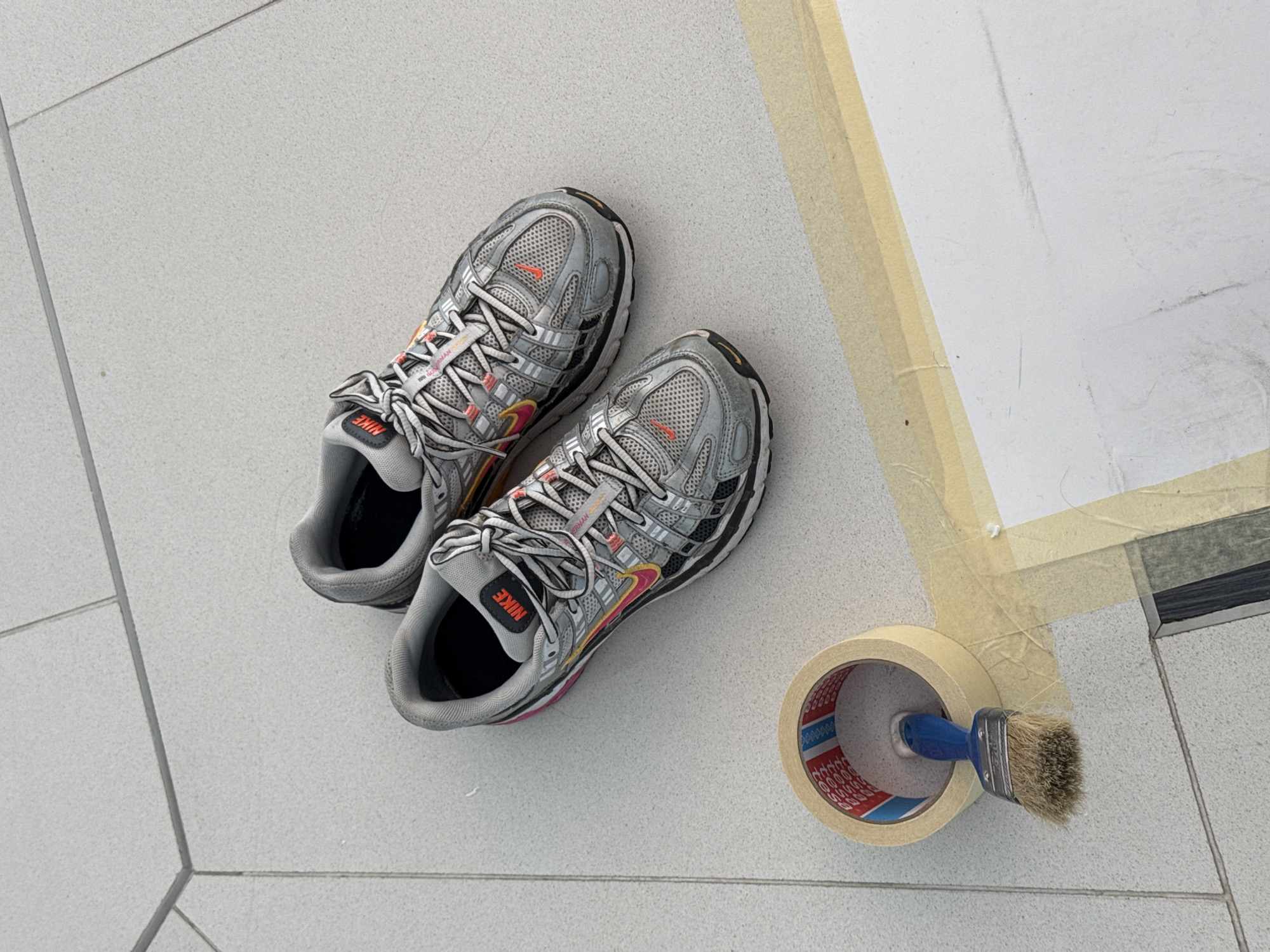 The scene shows a pair of grey Nike running shoes with red and yellow accents lying on a tiled floor, likely indoors. Next to them is a roll of masking tape with a blue-handled paintbrush stuck inside. The area near the shoes has been masked off with tape, suggesting painting is in progress. The tile pattern and overall aesthetic are consistent with modern interior design. The location is not discernible from the visual information. There are no people visible in the image.