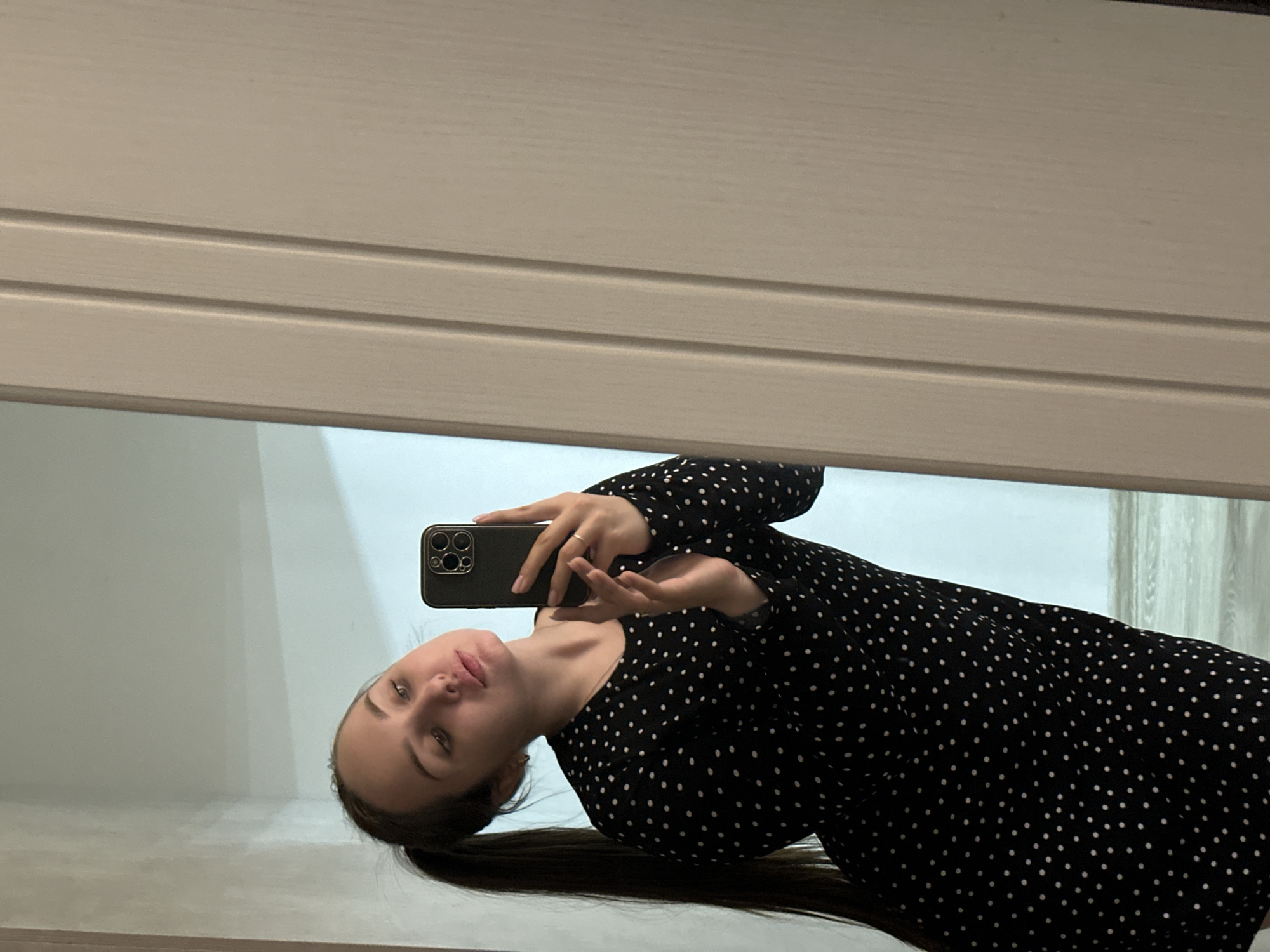The image captures a young woman taking a mirror selfie. The woman, with dark hair pulled back into a ponytail, looks directly at the camera with a neutral expression. She is wearing a long-sleeved black dress adorned with small white polka dots. In her right hand, visible in the reflection, she holds a dark-colored smartphone, likely an iPhone due to its distinctive multi-lens camera module.
The selfie is taken in a large mirror, which reflects her upper body and the phone. The physical background behind the mirror appears to be a light, possibly beige or pale wood, panel with horizontal grooves, possibly part of a closet door or furniture. The reflected background within the mirror shows a plain, light-colored wall, possibly off-white or very light blue, suggesting an indoor setting. There are no clear indications of time of day or weather, but the lighting suggests an interior space. No discernible text is visible on any surface or object. The setting is a room, possibly in Chaikovsky, Russia, as per the provided context.