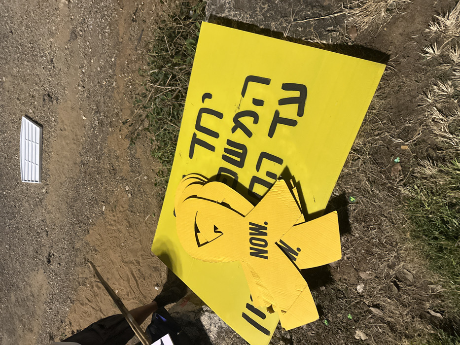 The image depicts a ground-level scene in Mishmar Hasharon, Israel, under artificial illumination at night. In the foreground, multiple yellow protest signs are visible on a dirt and gravel surface with sparse vegetation. A prominent rectangular yellow sign displays black Hebrew text that translates to "Together the mission until home." Stacked upon this sign are several yellow ribbon-shaped cutouts, each featuring the black English word "NOW."
To the left, the lower legs and feet of an individual are partially visible, wearing dark shorts and dark shoes. This individual holds two wooden sticks or poles diagonally across the frame. In the upper right background, a white rectangular object with horizontal slats, resembling a vent cover, rests on the bare ground. Shadows are cast by an unseen light source. No direct actions or interactions are currently depicted, but the arrangement of objects suggests preparation or aftermath related to a public display.