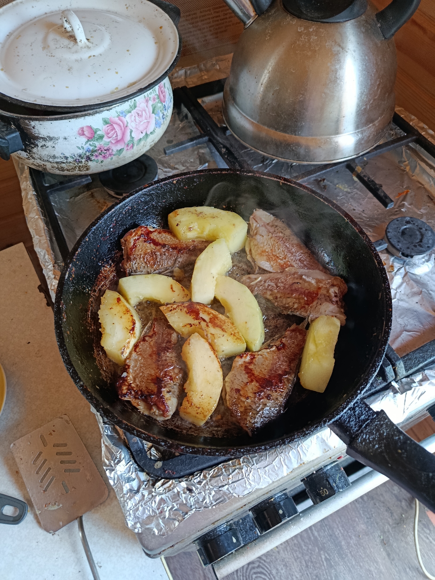 In a kitchen in Vareya, Russia, a black cast-iron frying pan is placed on a gas stove, cooking meat and potatoes. The pan, filled with sizzling food, is on the right side of the stove, with its handle extending towards the bottom right of the frame. To the left of the frying pan, a large, white enamel pot with a floral design of pink roses and blue flowers sits on another burner. Behind the pot, a silver kettle with a black handle is also on the stove. The stove top is covered in aluminum foil, and a beige spatula rests on a light-colored countertop to the left of the stove. The environment appears to be a home kitchen, with wooden walls visible in the background. The overall scene depicts a meal being prepared.