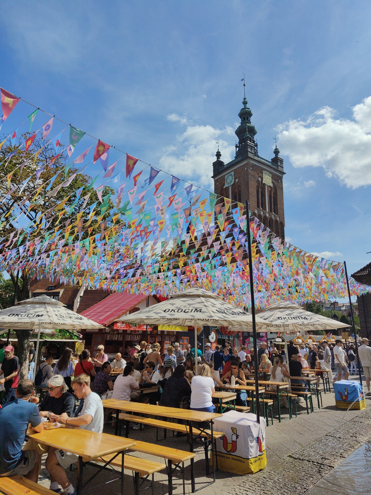 The image captures a lively outdoor scene on a sunny day in Gdańsk, Poland, likely in the historic Old Town area, given the prominent brick clock tower in the background which resembles the Main Town Hall. A multitude of colorful triangular flags are strung across the foreground, creating a festive canopy over a bustling outdoor seating area. Underneath large white umbrellas, branded with "OKOCIM," people are seated at long wooden tables and benches, engaged in eating, drinking, and conversing. The atmosphere appears relaxed and social, with a mix of adults and some children. In the background, more people mill about, suggesting a busy market or festival. The architecture visible behind the seating area includes buildings with red tiled roofs and traditional facades, further grounding the scene in a European city setting. The ground is paved, and a water feature, possibly a fountain or canal, is visible on the right side of the image, reflecting the light.