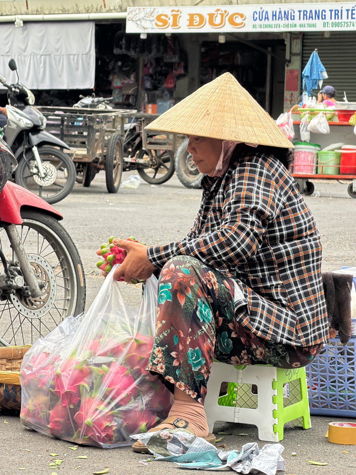 The image depicts a street vendor in Nha Trang, Vietnam, selling dragon fruit. The vendor, wearing a traditional conical hat and a plaid shirt over floral-patterned pants, is seated on a green and white folding stool. They are holding a bunch of dragon fruit, with a large clear plastic bag filled with more fruit beside them. In the background, a store sign with Vietnamese text, "CỬA HÀNG TRANG TRÍ TẾT", is visible, indicating it's a decoration store, likely for the Lunar New Year. Several motorbikes and a multi-wheeled cart are also present in the street, suggesting a busy market or roadside selling environment. The ground is paved, with some green leaves scattered around the vendor.