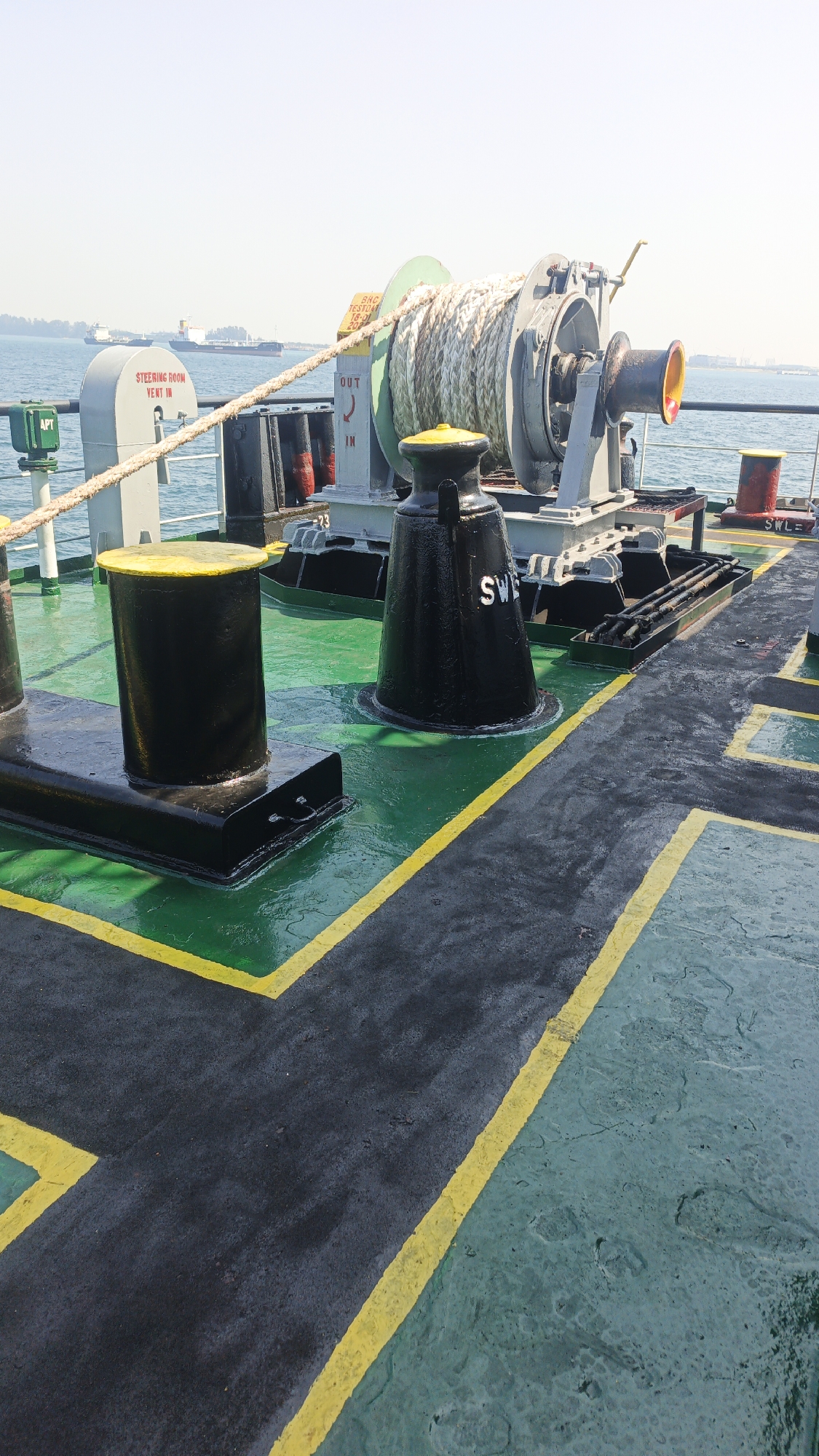 The image shows a deck of a ship, likely a cargo vessel, with a large winch and mooring equipment. The deck is painted green and black with yellow safety lines. The winch is made of metal and has a large drum with thick rope wound around it. There are also two black bollards with yellow tops, used for securing mooring lines. In the background, there is the sea and other ships. A sign on the winch reads "OUT IN". Another sign on a grey structure to the left reads "STEERING ROOM VENT IN". The image appears to have been taken during the day, with clear skies and bright sunlight. The overall impression is that of a functional, working ship at sea. There are no people visible in the image. The scene is likely within Singapore's busy port, given the presence of multiple vessels and the overall industrial maritime environment.