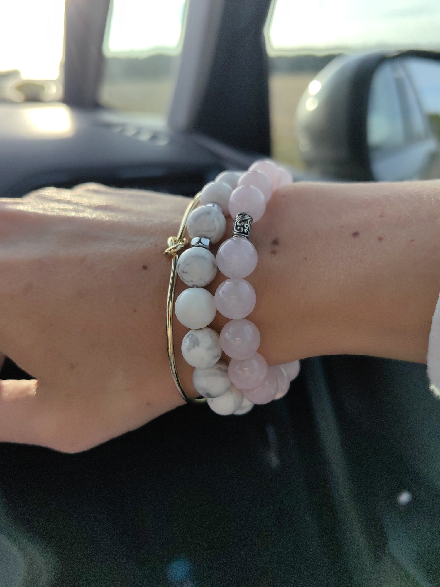 A close-up shot focuses on a person's arm wearing two beaded bracelets. The bracelet closer to the camera is gold-toned and appears to be a bangle style with a knot detail. It is adorned with round, white beads with grey marbling, interspersed with a few smooth white beads. The second bracelet, layered on top, is made of larger, smooth, pale pink beads. Both bracelets are on the person's left wrist. The skin is lightly tanned and shows a few small dark marks, possibly moles or freckles. The background is blurred, suggesting the shot was taken from inside a vehicle. Sunlight streams in from the left, creating a bright, hazy glow and casting a sheen on the arm. The blurry background hints at the interior of a car, with what appears to be a dashboard or console in the lower part of the frame, and a side-view mirror or window reflection visible to the right. The image does not provide any information or context related to the city of Ilyanskiy selskiy Sovet, Belarus.