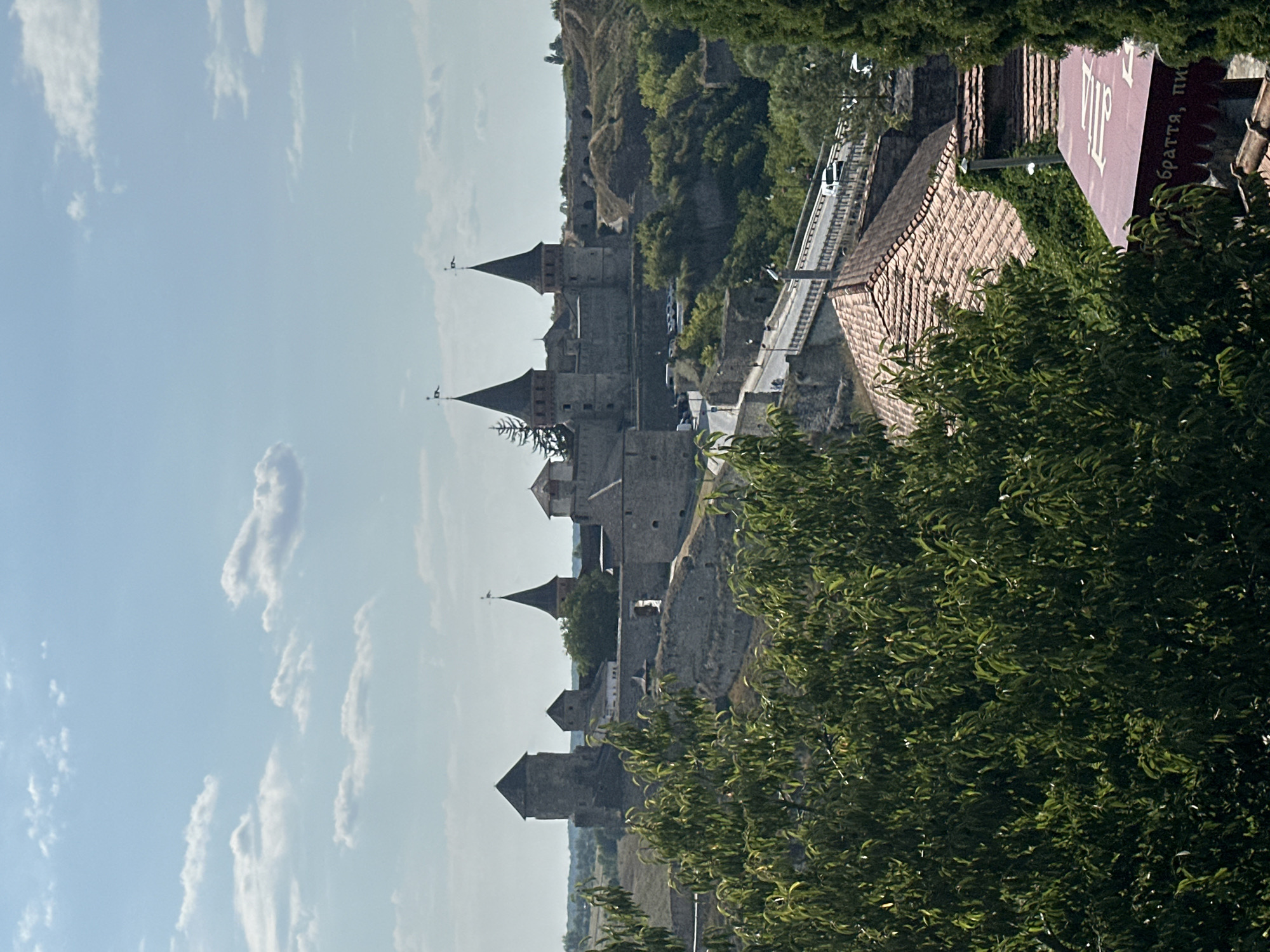 The image depicts a sunny day with scattered clouds over the Kamianets-Podilskyi Castle in Ukraine. The castle, a large stone fortress with conical towers, is situated on a hill, surrounded by greenery. In the foreground, lush green trees partially obscure the view. A road with a bridge crosses in front of the castle, with vehicles visible on it. The architecture of the castle suggests historical significance. The overall scene is one of a picturesque and historic landmark under a clear sky.