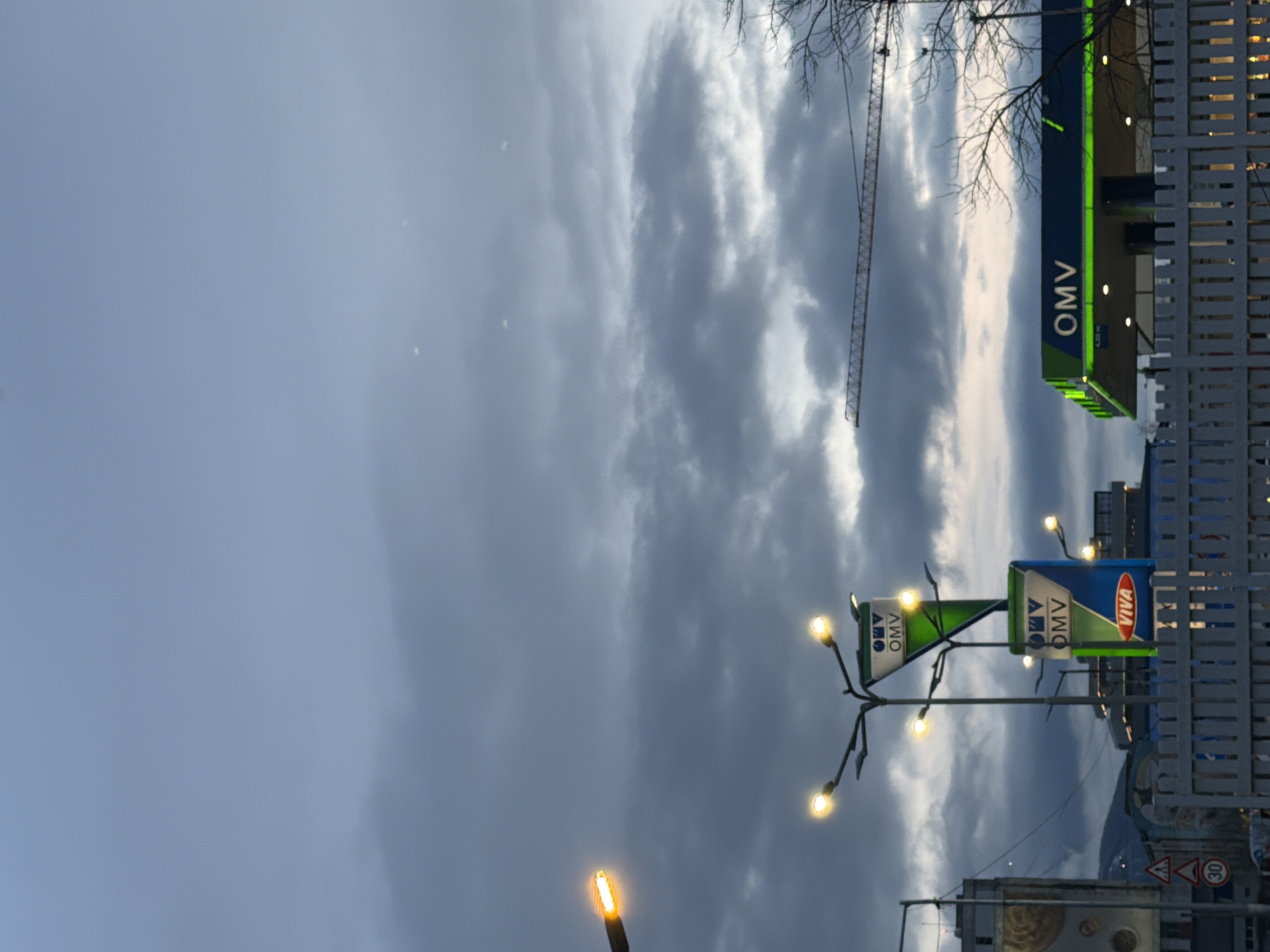 The image shows an OMV petrol station in Sofia, Bulgaria. The sky is overcast. A construction crane is visible in the background. Street lights illuminate the area. The OMV logo and branding are prominently displayed. There are also road signs visible.