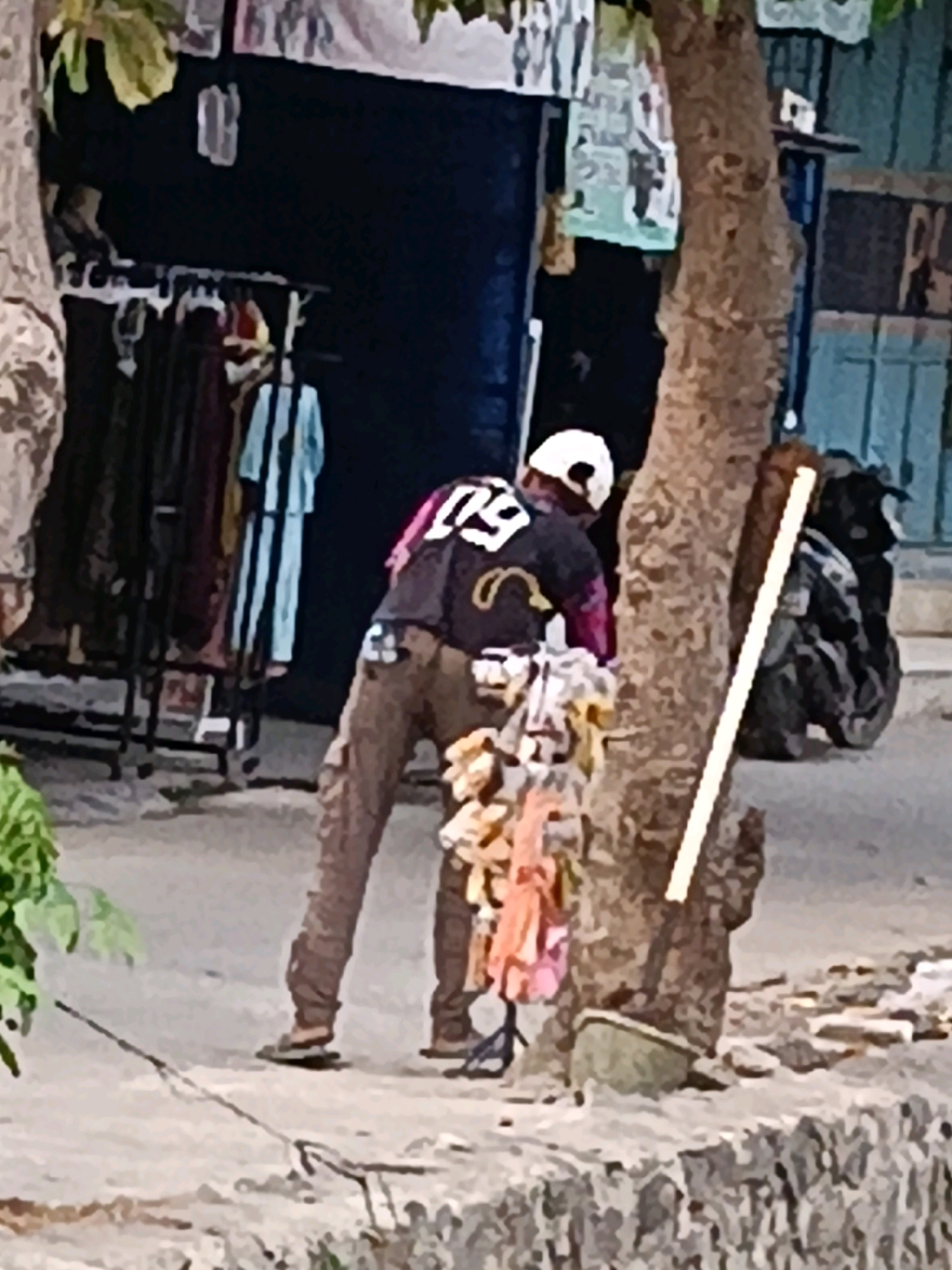 A person wearing a white cap and dark clothing with a white numerical design on the back is standing by a tree. They are holding a collection of colorful items. Behind them, clothing is displayed on racks, and a motorcycle is parked. The scene appears to be an outdoor marketplace in Belimbing, Indonesia.