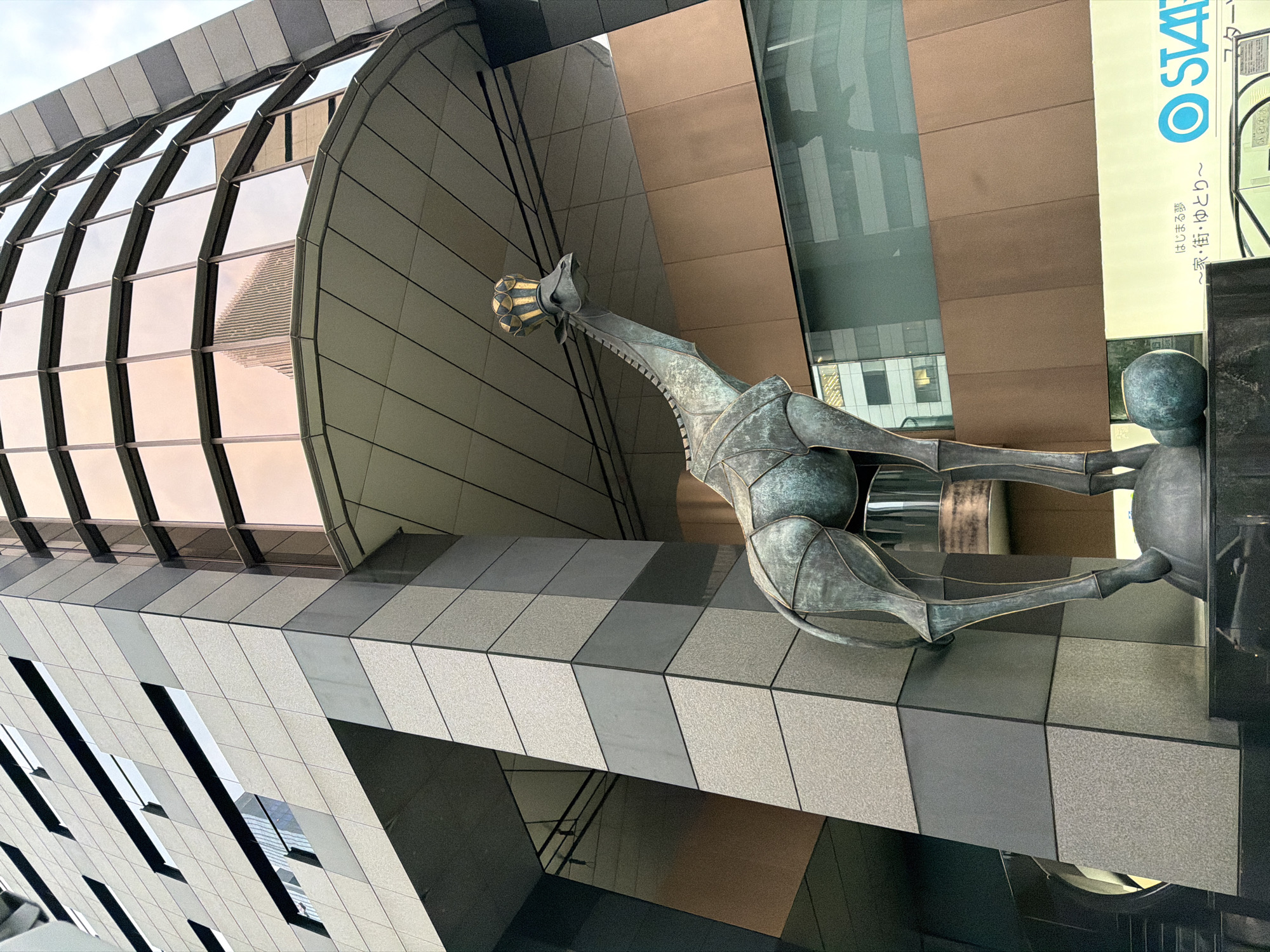 An outdoor, eye-level, medium shot captures a bronze sculpture of a giraffe wearing a golden crown, positioned in front of a modern, multi-story building with copper-tinted windows. The giraffe sculpture is detailed with segmented panels, giving it a stylized, almost robotic appearance. It stands on a dark pedestal with two rounded spheres at its base. The building features a facade of textured gray panels and large, reflective windows that mirror other buildings. The sky is overcast. Part of a business name with Japanese characters and a blue "STARTS" logo are visible at the bottom right of the image, indicating the location is likely within a commercial area in Chuo, Japan.