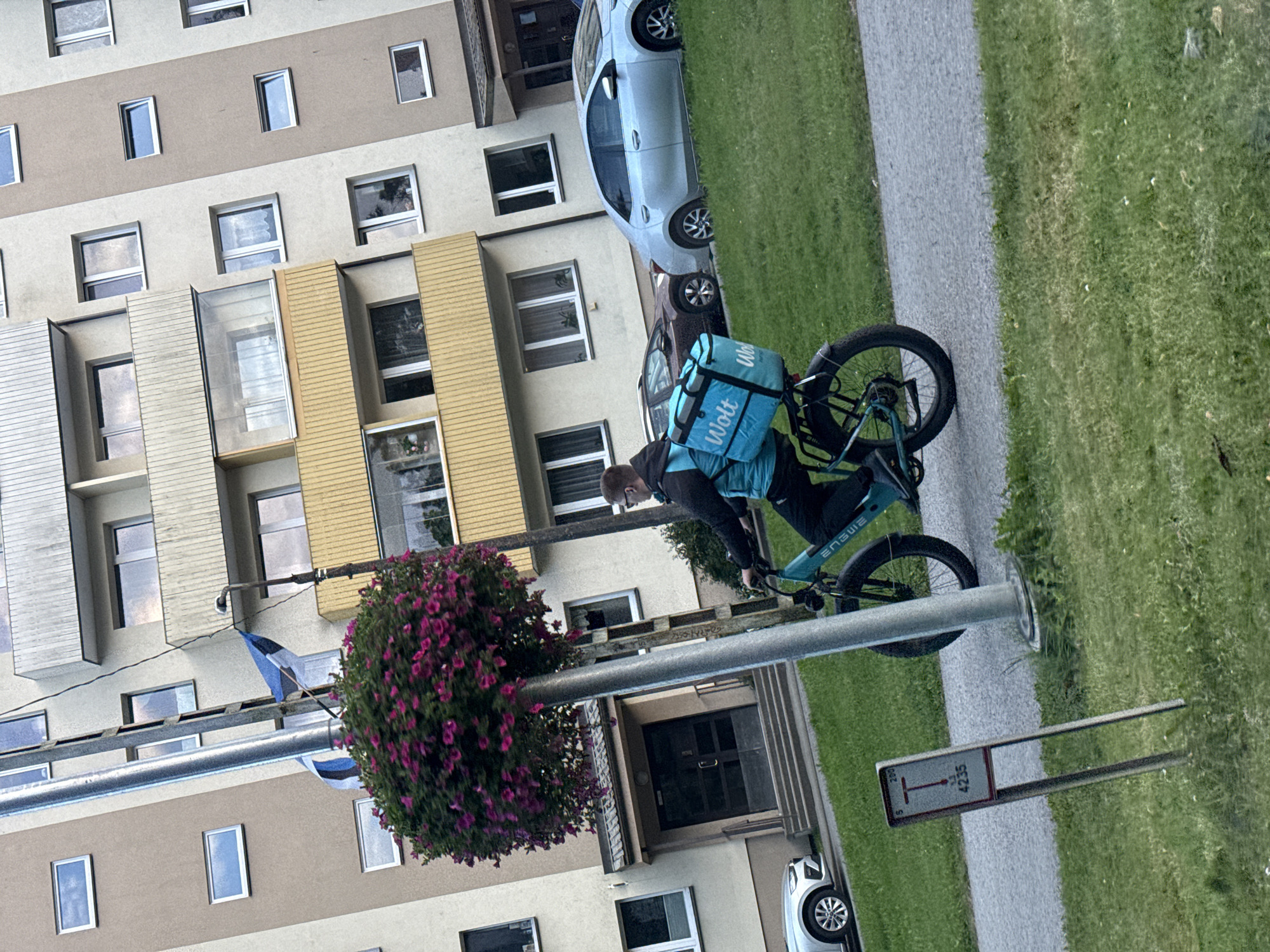 A person wearing a blue vest with the Wolt logo rides a teal bicycle with a large blue insulated bag on the back. The person is wearing dark pants and a light grey shirt. They are on a paved path next to a grassy area. In the background, there is a multi-story apartment building with beige and brown siding and yellow accents. A flag is visible on a pole attached to the building. A silver car is parked on the left side of the image, and a darker car is parked behind it. A small signpost with the letter "T" and the number "4235" is visible in the grass. The scene appears to be in a residential area of Tallinn, Estonia.