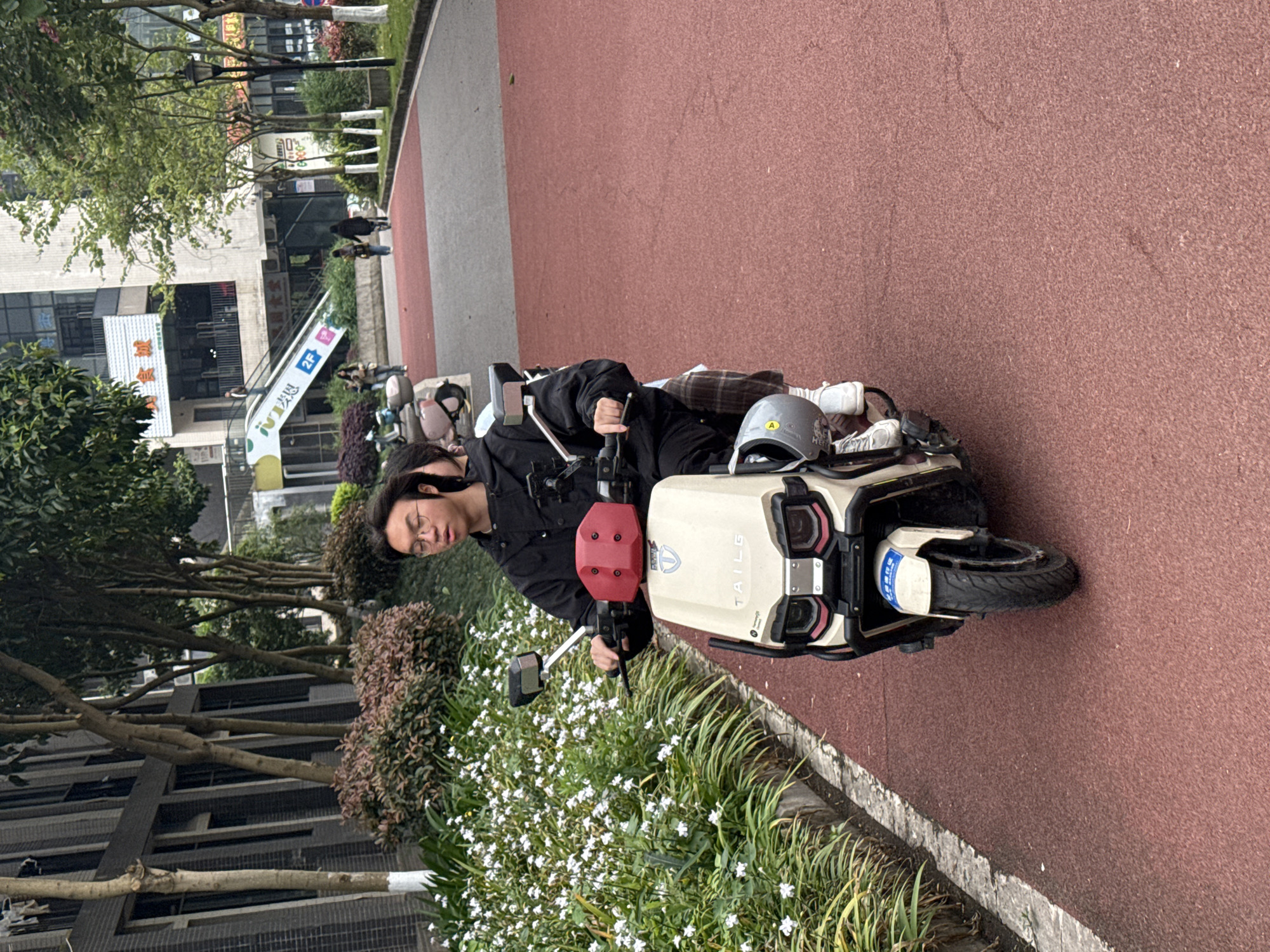 A person with glasses is sitting on a white electric scooter. There is a grey helmet on the scooter. They are on a red paved path. The buildings in the background have Chinese writing on them. The location is Jiangjin, China.