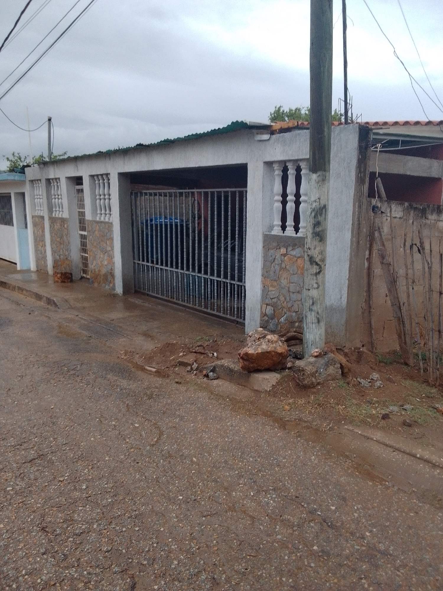 The image depicts a street scene in Juan Griego, Venezuela, characterized by overcast skies and a generally unkempt environment. In the foreground, a rough asphalt road shows signs of wear and tear, with scattered gravel and debris. To the right of the road, a dirt embankment is visible, featuring loose soil and rocks.
Dominating the midground is the facade of a building with a white stucco finish, adorned with decorative stone accents and vertical railings. A metal gate, likely a driveway entrance, is prominent, with what appear to be two large blue bins visible behind it. A concrete pillar, weathered and marked with graffiti, stands near the gate. Adjacent to the gate is a wall constructed from wooden planks supported by rustic framing, showing signs of disrepair. A large, reddish-brown boulder rests on the ground near the gate and pillar.
The background reveals more of the building's structure, including a corrugated green roof and segments of a tiled roof. Overhead, electrical wires crisscross the sky, supported by a wooden utility pole. Trees are visible behind the building to the left. The overall impression is of a residential or commercial area in a state of modest upkeep, with evidence of ongoing use and weathering.