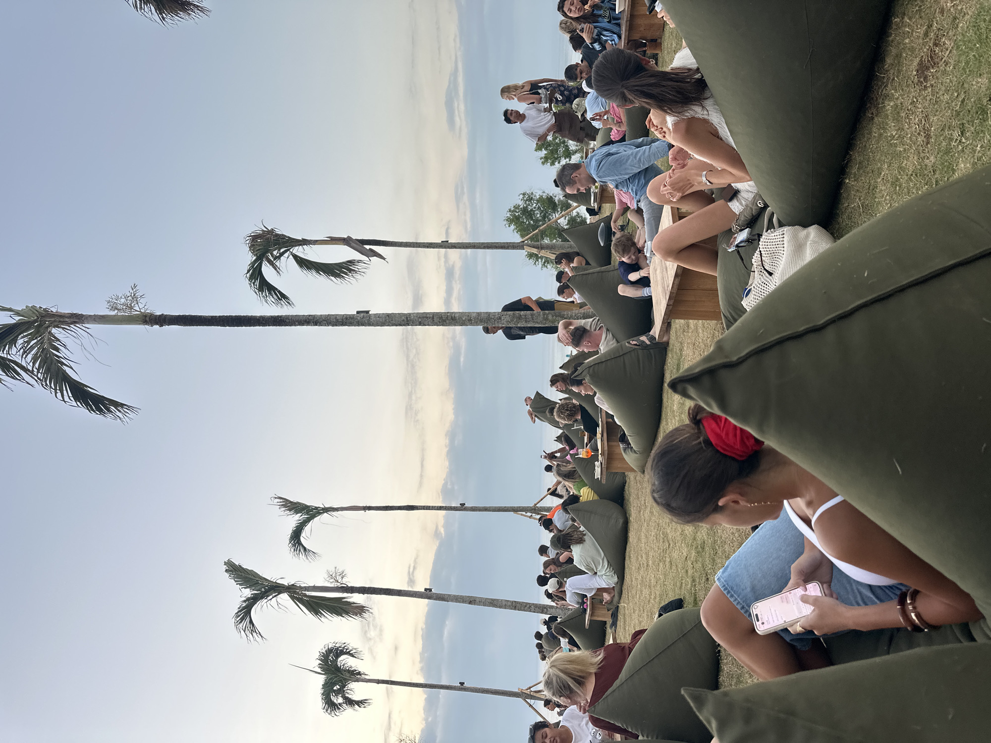 In Kuta Selatan, Indonesia, an outdoor gathering takes place on a grassy lawn, facing a body of water. A multitude of people are seated comfortably on large, olive-green beanbag chairs, many engaged in casual conversation, some looking at their phones, and others enjoying drinks. In the foreground, a woman in a white top and denim shorts is looking at her phone. Behind her, other individuals are similarly relaxed on beanbags. Tall palm trees punctuate the scene, with the sky displaying a soft gradient of blues and grays, suggesting either sunrise or sunset. The overall atmosphere is one of leisure and community.