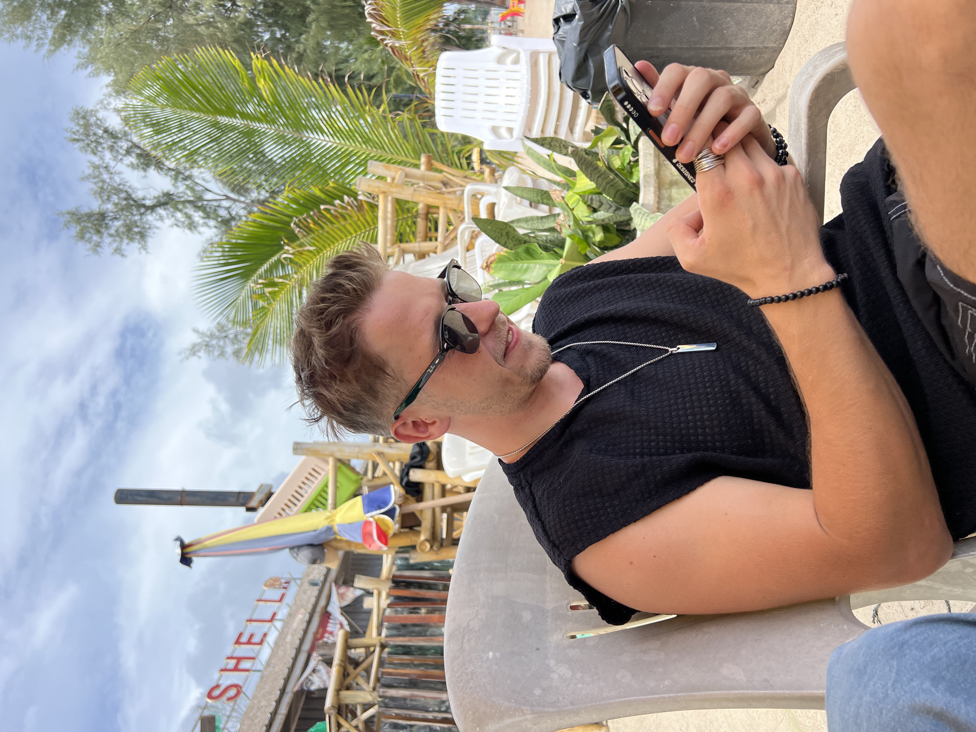 A young Caucasian man with blond hair and sunglasses sits in a light gray plastic chair, looking down at his smartphone. He is wearing a black sleeveless shirt, a silver necklace with a rectangular pendant, and a black beaded bracelet on his left wrist. He also wears several silver rings on his right hand. The background shows a beach scene with palm trees, bamboo structures, colorful umbrellas, and stacked white plastic chairs. The sky is cloudy. The man is holding the phone horizontally and appears to be typing or scrolling.