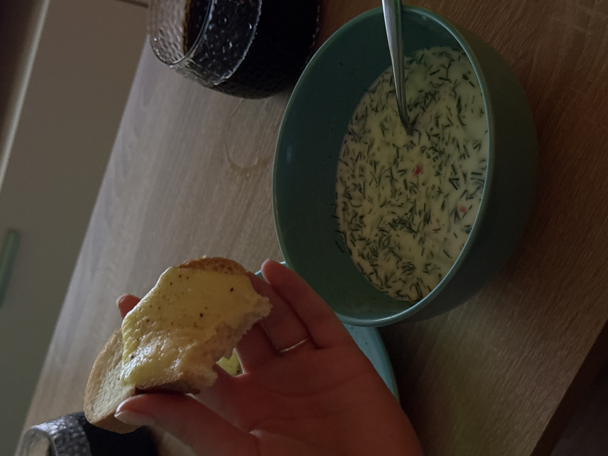 In a close-up shot, a hand holds a slice of toasted bread topped with a yellow spread, possibly cheese or butter, seasoned with black pepper. The bread is slightly torn, revealing a fluffy interior. A thin silver ring adorns a finger on the hand. In the background, a teal bowl contains a creamy white soup or dip with finely chopped green herbs, likely dill, and possibly some red specks. A silver spoon rests in the bowl. To the right, a dark beverage fills a textured glass. The setting appears to be a wooden table, with a hint of a blurred background suggesting an indoor environment.