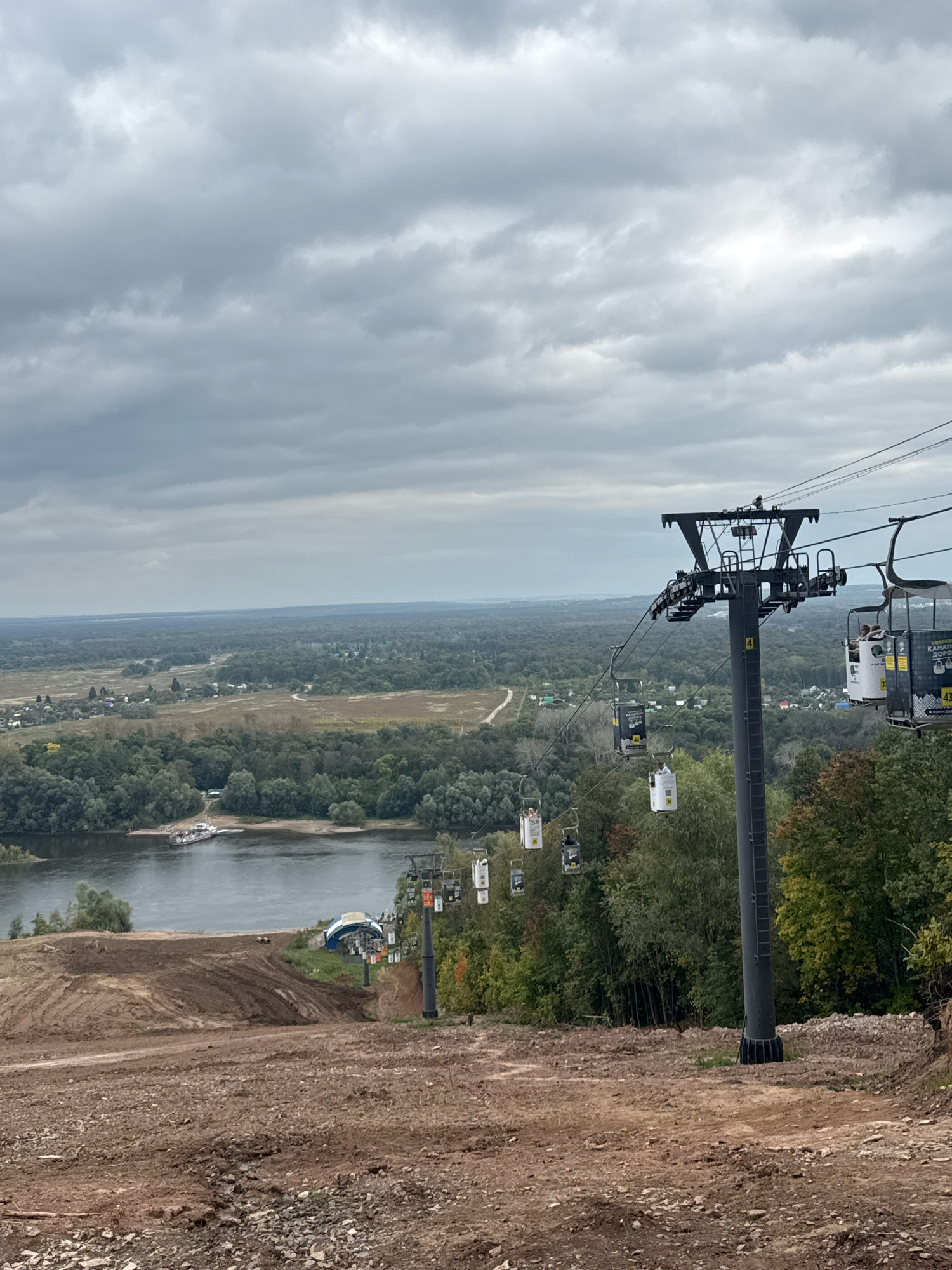 An elevated view captures a ski lift with multiple gondolas ascending a steep, partially constructed slope of bare earth and rocks. The ski lift tower, a tall dark gray structure, stands prominently on the right side of the frame, supporting the cables. Several gondolas, white and dark colored, are visible suspended from the cables, with a few containing passengers. The slope descends towards a wide river, with a boat docked on its bank. Beyond the river, a dense green forest stretches into the distance, dotted with a small settlement of houses. The sky above is overcast with thick, gray clouds. The scene is located at the "Ufa-Arena" ski resort in Ufa, Russia, a site known for its recreational facilities.