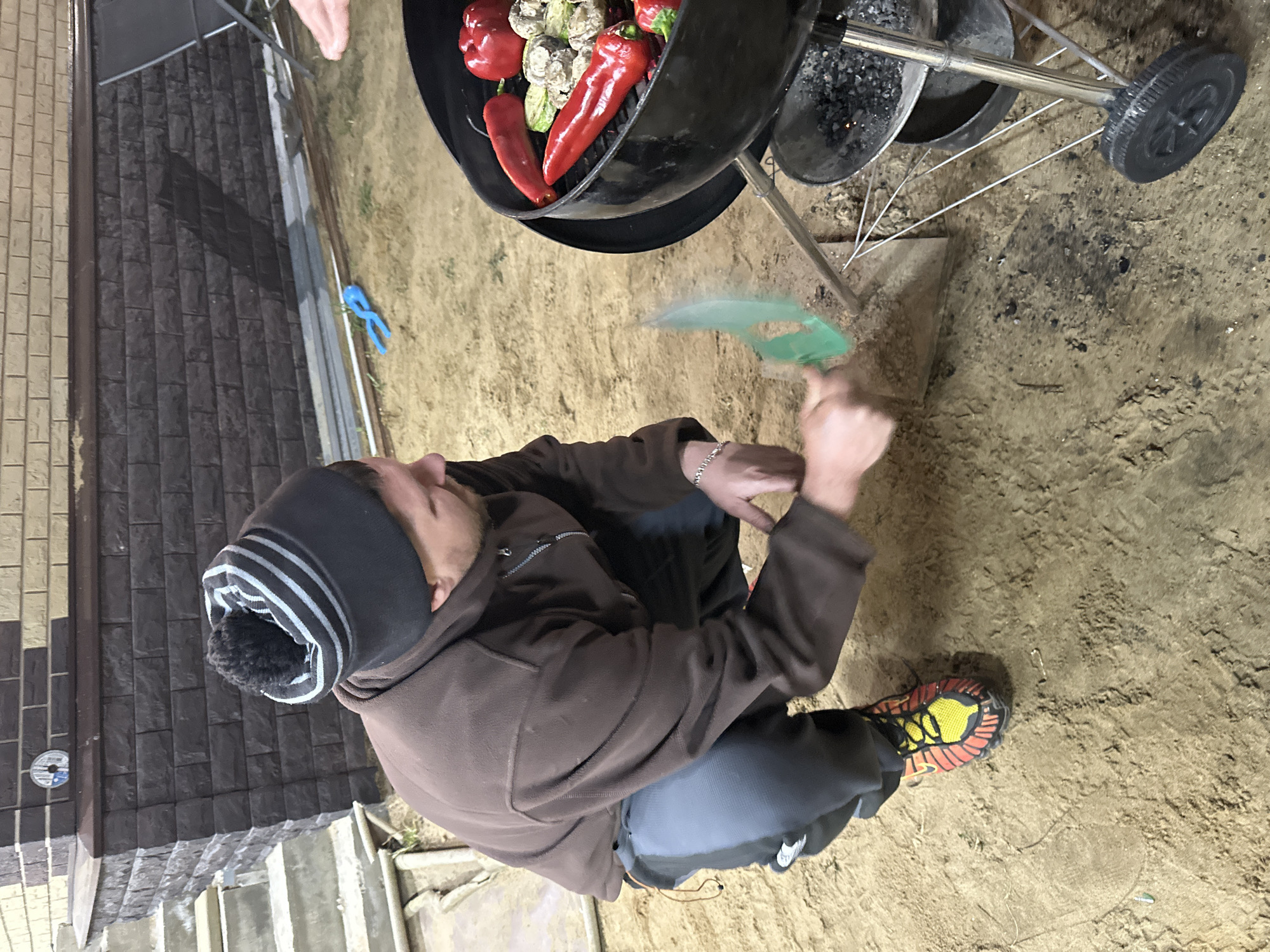 A man wearing a brown hooded jacket, black pants, and a gray and white beanie is crouched down and fanning a charcoal grill. The grill contains red bell peppers and small white vegetables, potentially mushrooms. The man is using a green fan to ignite the charcoal. He is wearing orange and black shoes. The ground appears to be dirt or sand. Part of a wall constructed of brick-like materials is visible in the background to the left. A pair of blue pliers are present on the ground behind the grill. The location appears to be outside, likely in слобода Старая, Russia.