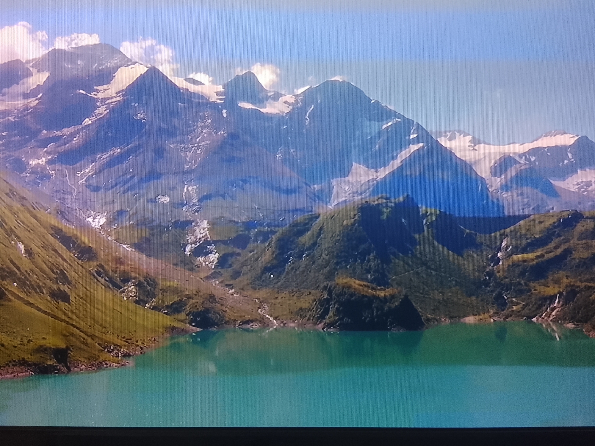 The image depicts a serene mountain landscape with a body of turquoise water in the foreground. Towering, snow-capped mountains dominate the background, their peaks piercing a clear blue sky. Lush green slopes descend from the mountains towards the water, dotted with rocky outcrops and patches of snow. The water itself is calm and reflects the surrounding scenery, with subtle ripples on its surface. There are no people or man-made structures visible in the image.
This image does not depict the city of Ivanovo, Russia, or any location within it. The scene is clearly a natural mountain environment, inconsistent with the urban and industrial characteristics of Ivanovo.