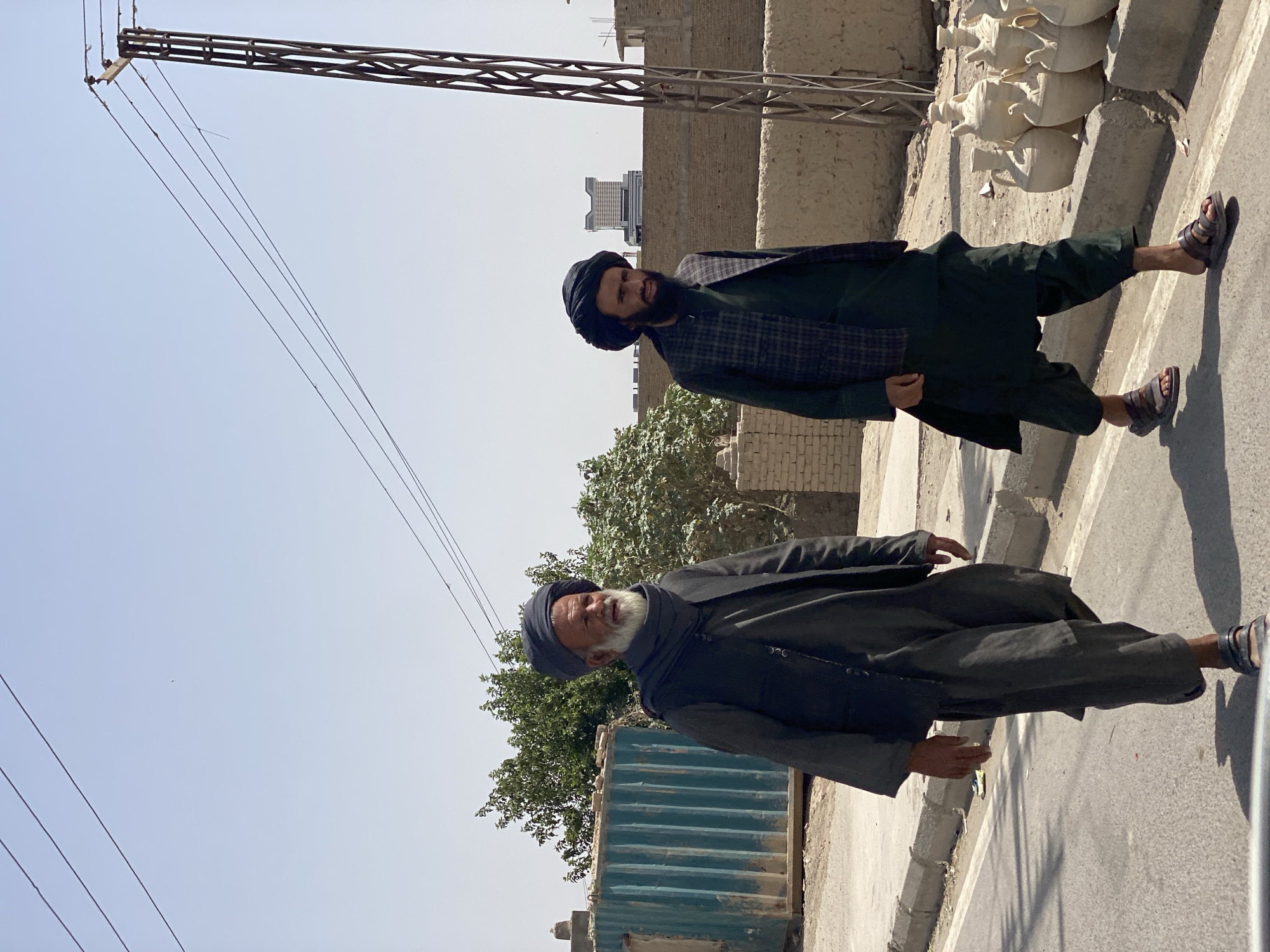 Two men walk on a dusty road in Kandahar, Afghanistan. The man on the left is older, with a white beard and a dark turban. He wears a dark vest over a long dark robe and loose pants. The man on the right has a beard and wears a dark turban and a plaid vest over a dark robe and pants. Both men wear sandals. In the background, there is a teal shipping container, some brick walls, a utility pole with wires, and a modern building in the distance. A collection of clay pots is visible on a ledge to the right. The sky is clear and blue.