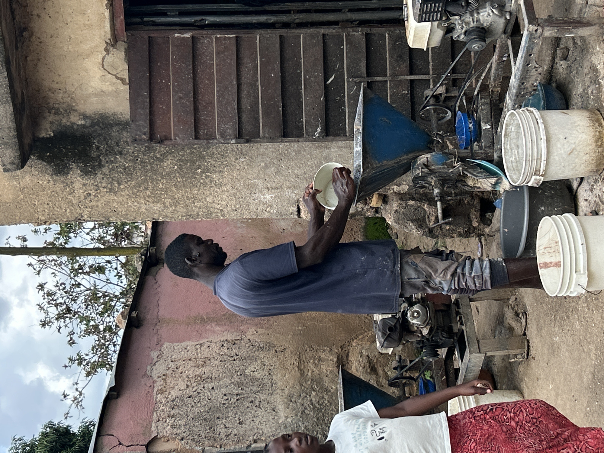 In Jos, Nigeria, a man in a dark blue t-shirt and patterned pants is pouring a white substance from a bowl into a large blue funnel of an industrial grinding machine. To his left, a young girl in a white t-shirt and a red patterned skirt stands looking towards the left of the frame. The scene appears to be an outdoor workshop or processing area, with various pieces of machinery, buckets, and building structures visible. The ground is dirt and concrete. The buildings in the background are made of textured concrete and painted pink.