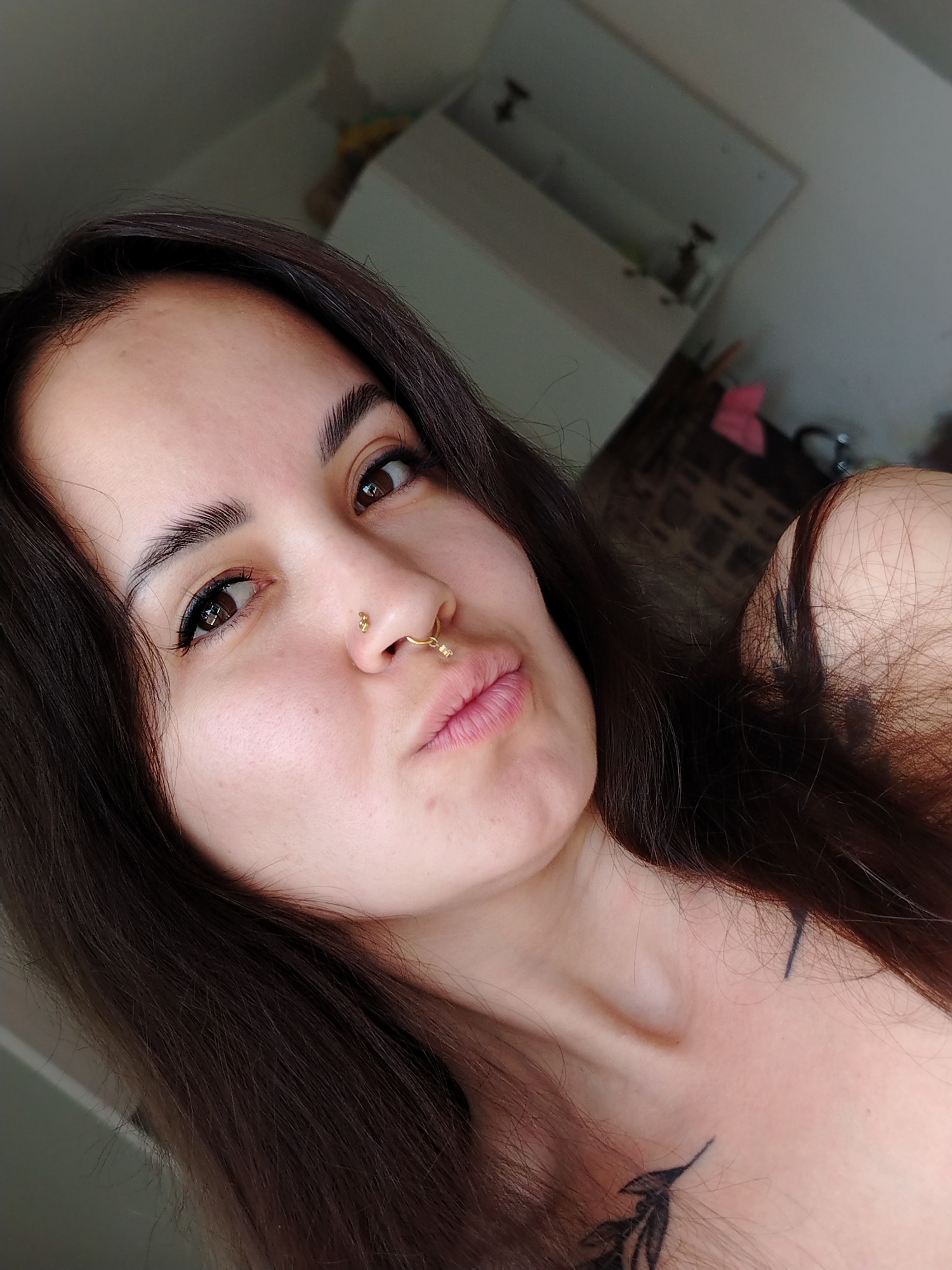 A close-up selfie depicts a young woman with long, dark brown hair and fair skin. She has dark eyes, full eyebrows, and is wearing a gold nose ring and stud. Her lips are slightly puckered. Two small tattoos are visible on her neck and upper chest. The background is slightly blurred and shows a white cabinet, a brick-patterned wall, and what appears to be a sink and faucet. The location, Noviy, Russia, is extraneous as the image itself does not provide specific markers of location.