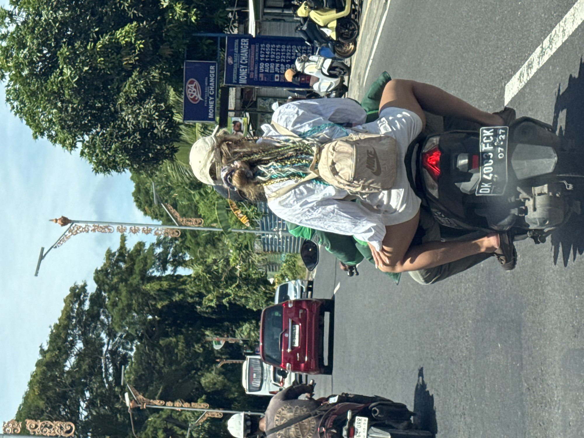 A partially obscured view reveals a street scene in Nusa Dua, Indonesia. The image is taken from a driver's perspective, likely inside a vehicle. A motor scooter carries two individuals: a driver wearing green and a passenger with tan backpack, a white cap, and what appears to be dreadlocks, is riding pillion. The scooter's license plate reads "DK 4063 FCK" and its registration appears to expire "07-27". The passenger is wearing brown sandals. The vehicle is on a paved road marked with solid white lines. The background features a "MONEY CHANGER" sign, a yellow motor scooter, a red car, trees, and decorative street lighting. The driver's shadow is visible on the road. Other vehicles are visible in the road.